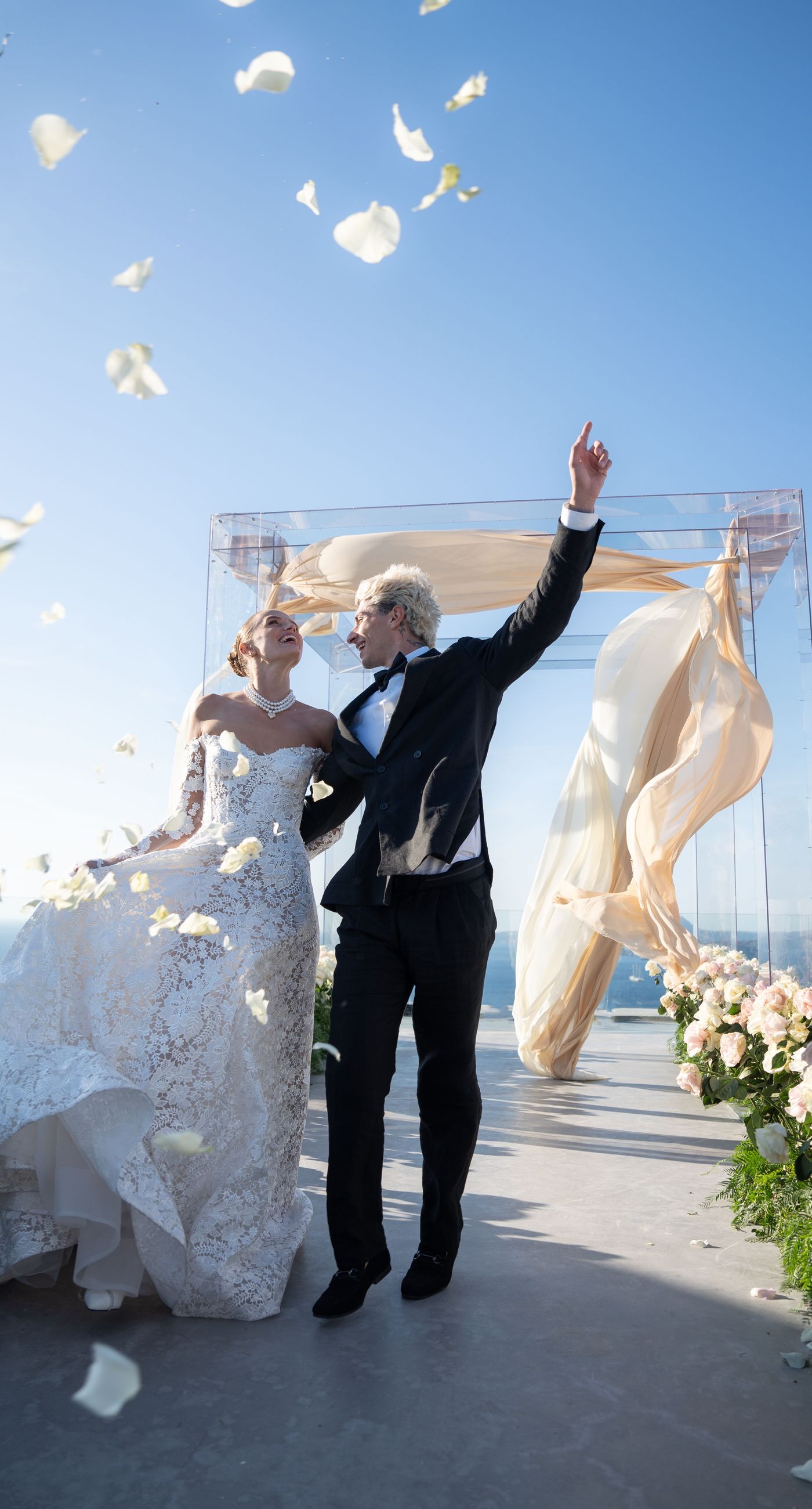 Isabella & Oliver - Under a clear blue sky, what unfolds is an ethereal expression of elegance and romance. The serene backdrop of vast, azure waters beautifully contrasts with the soft, delicate whites of the cascading petals, creating a scene both harmonious and enchanting. Meticulously arranged roses in gentle pinks and creams line the path, complementing the timeless lace and flowing structure of the bride's gown, while the groom's classic black suit adds a touch of refined sophistication. The graceful drapery, kissed by the gentle breeze, frames this moment of joy and forms a masterpiece of floral artistry, balancing nature's serenity with bridal grandeur. - Photo 2