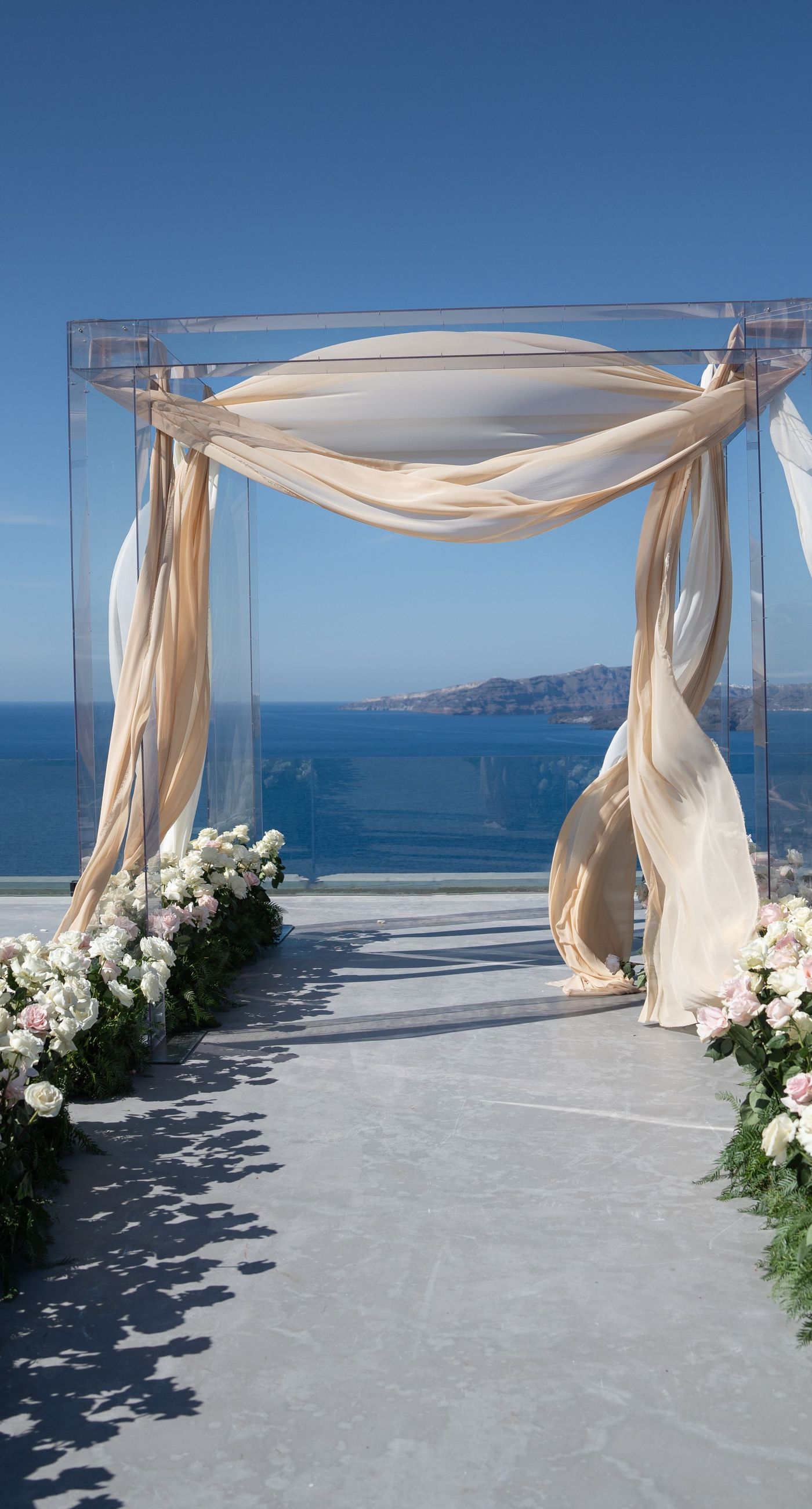 Isabella & Oliver - Under a clear blue sky, what unfolds is an ethereal expression of elegance and romance. The serene backdrop of vast, azure waters beautifully contrasts with the soft, delicate whites of the cascading petals, creating a scene both harmonious and enchanting. Meticulously arranged roses in gentle pinks and creams line the path, complementing the timeless lace and flowing structure of the bride's gown, while the groom's classic black suit adds a touch of refined sophistication. The graceful drapery, kissed by the gentle breeze, frames this moment of joy and forms a masterpiece of floral artistry, balancing nature's serenity with bridal grandeur. - Photo 1