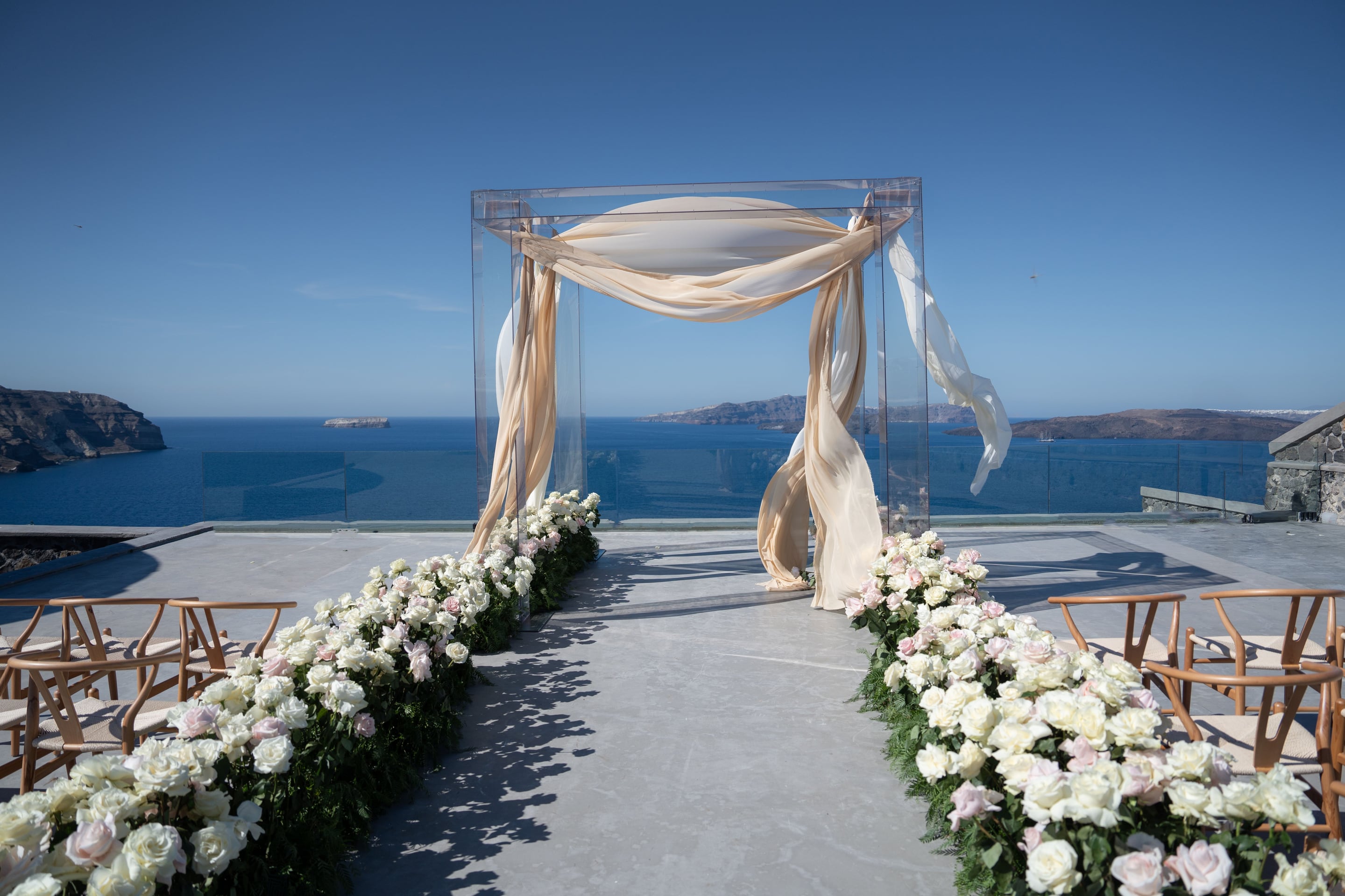 A clear acrylic wedding arch draped with cream and champagne silk chiffon stands at the end of an aisle on a pale stone terrace, with the Aegean and the caldera islands stretching blue to the horizon beyond a glass balustrade. Both sides of the aisle are lined with low runners of white garden roses blushed with pale pink, tucked into dark green foliage, and wooden wishbone chairs wait on the right. This is a wedding ceremony set at Cavo Ventus in Santorini, Greece, under a clear sky.