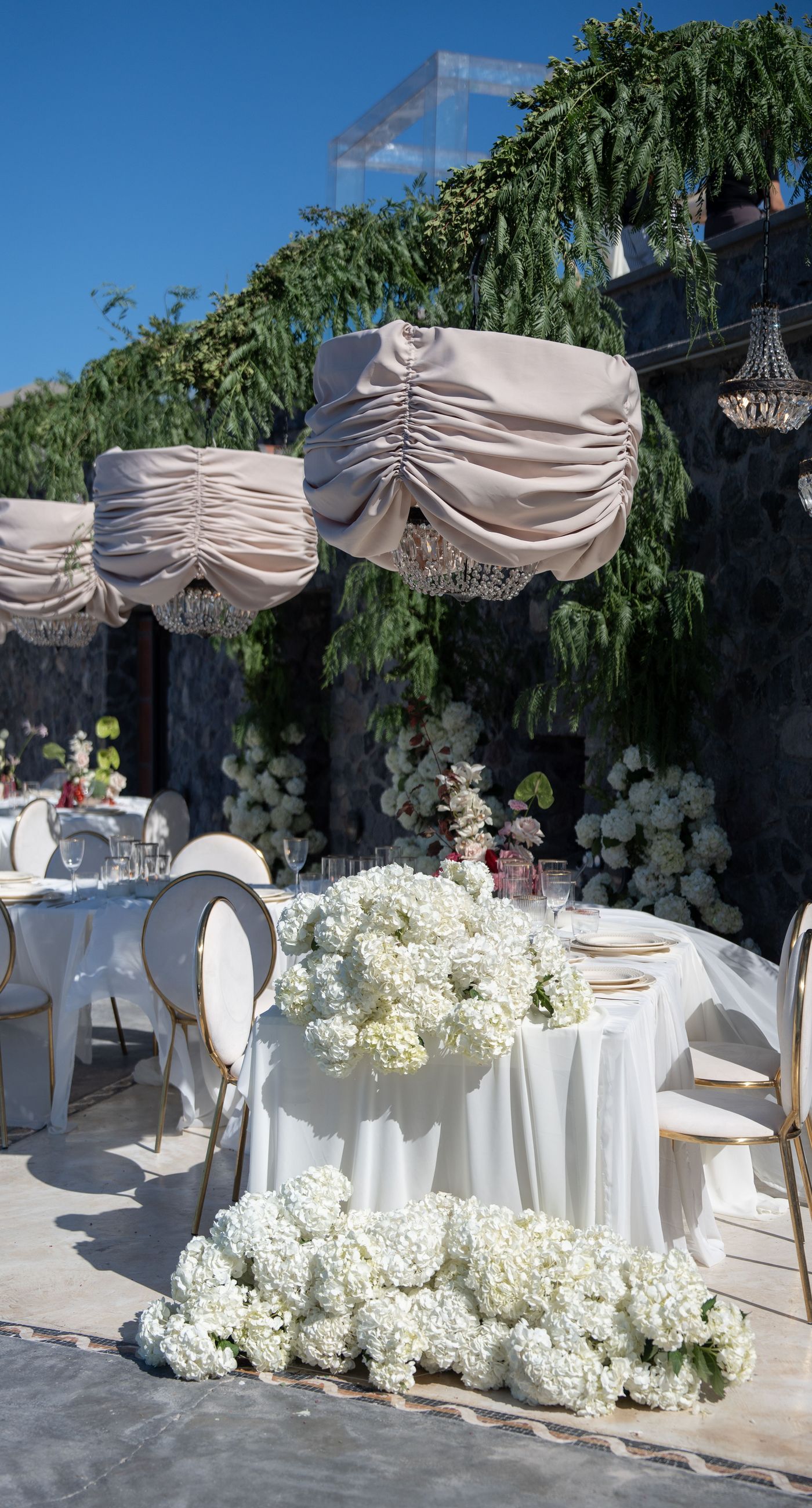 Isabella & Oliver - Under a clear blue sky, what unfolds is an ethereal expression of elegance and romance. The serene backdrop of vast, azure waters beautifully contrasts with the soft, delicate whites of the cascading petals, creating a scene both harmonious and enchanting. Meticulously arranged roses in gentle pinks and creams line the path, complementing the timeless lace and flowing structure of the bride's gown, while the groom's classic black suit adds a touch of refined sophistication. The graceful drapery, kissed by the gentle breeze, frames this moment of joy and forms a masterpiece of floral artistry, balancing nature's serenity with bridal grandeur. - Photo 16