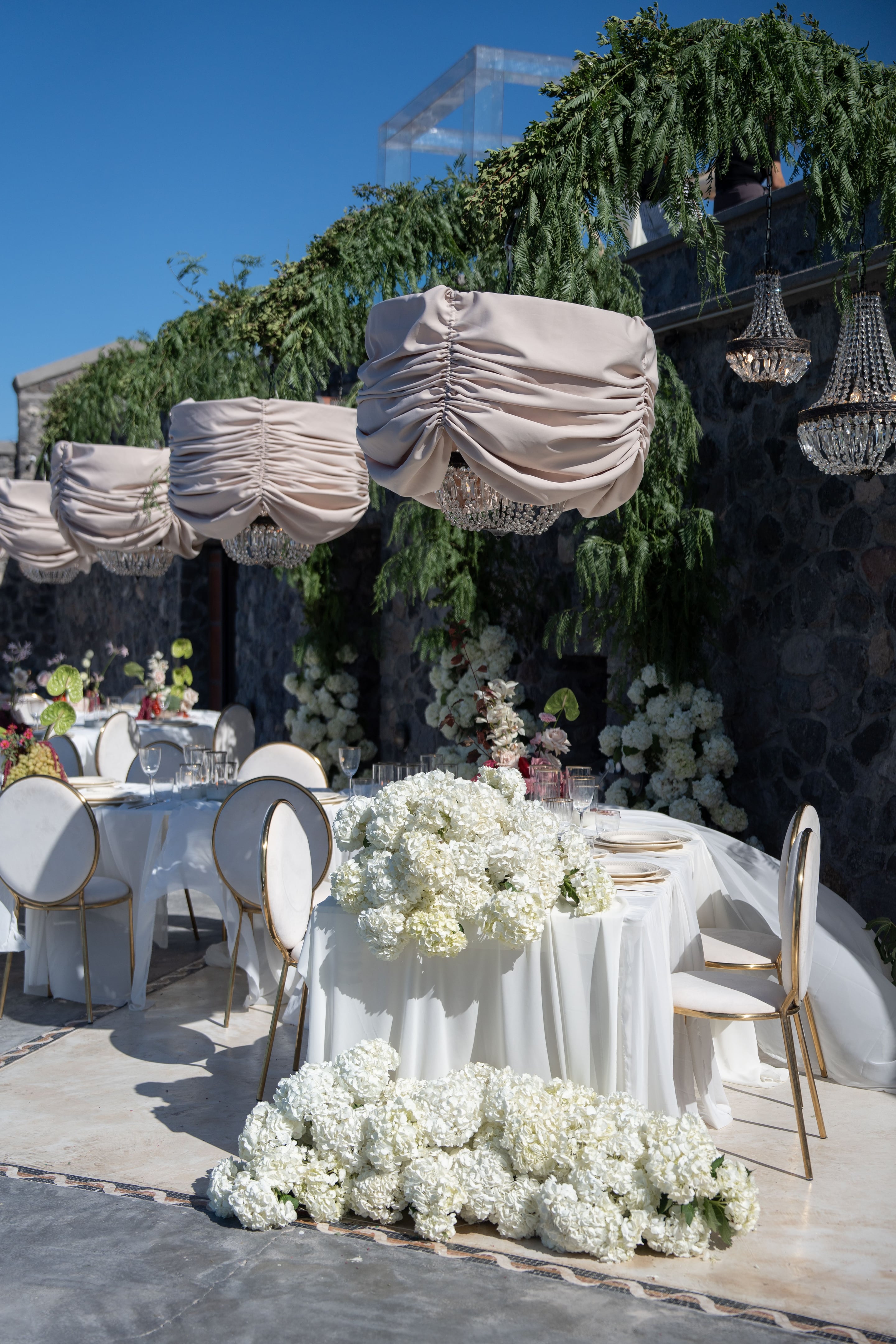 Generous clusters of white hydrangeas spill across a long, linen-draped wedding table at Cavo Ventus in Greece, with more blooms tumbling down in front of the stone wall. Gold-rimmed cream chairs and pale pink fabric gathered beneath crystal chandeliers frame the scene, while sprays of white flowers and trailing green fern rise against the dark stone. The soft, sunlit terrace makes it feel airy and romantic.