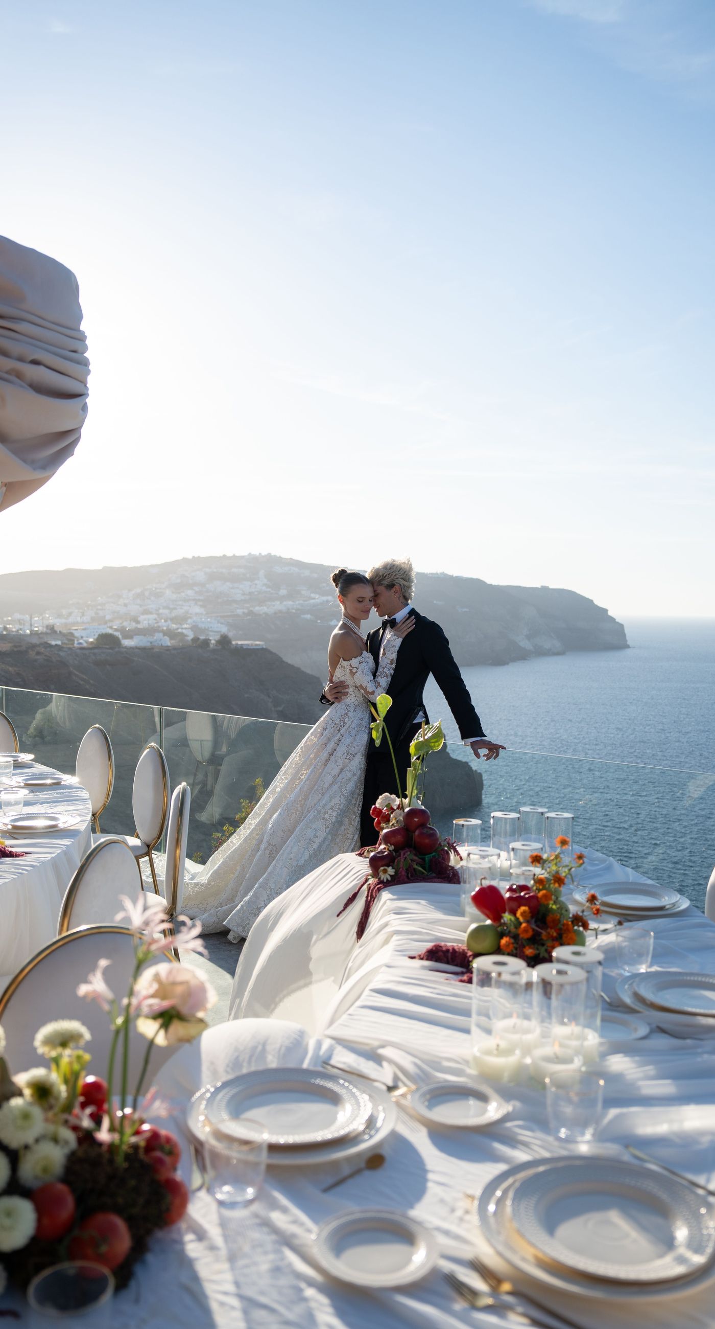 Isabella & Oliver - Under a clear blue sky, what unfolds is an ethereal expression of elegance and romance. The serene backdrop of vast, azure waters beautifully contrasts with the soft, delicate whites of the cascading petals, creating a scene both harmonious and enchanting. Meticulously arranged roses in gentle pinks and creams line the path, complementing the timeless lace and flowing structure of the bride's gown, while the groom's classic black suit adds a touch of refined sophistication. The graceful drapery, kissed by the gentle breeze, frames this moment of joy and forms a masterpiece of floral artistry, balancing nature's serenity with bridal grandeur. - Photo 7
