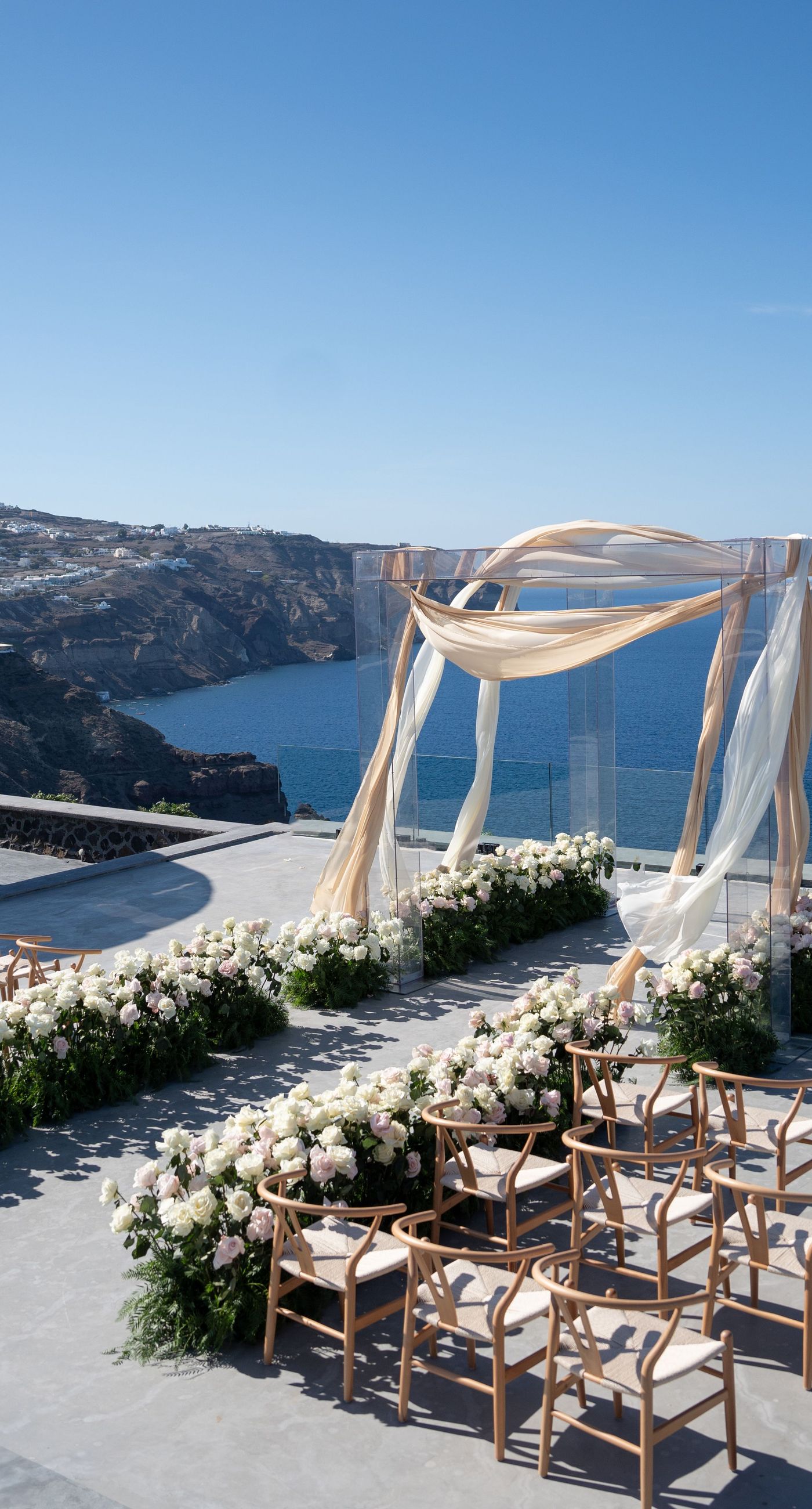 Isabella & Oliver - Under a clear blue sky, what unfolds is an ethereal expression of elegance and romance. The serene backdrop of vast, azure waters beautifully contrasts with the soft, delicate whites of the cascading petals, creating a scene both harmonious and enchanting. Meticulously arranged roses in gentle pinks and creams line the path, complementing the timeless lace and flowing structure of the bride's gown, while the groom's classic black suit adds a touch of refined sophistication. The graceful drapery, kissed by the gentle breeze, frames this moment of joy and forms a masterpiece of floral artistry, balancing nature's serenity with bridal grandeur. - Photo 3