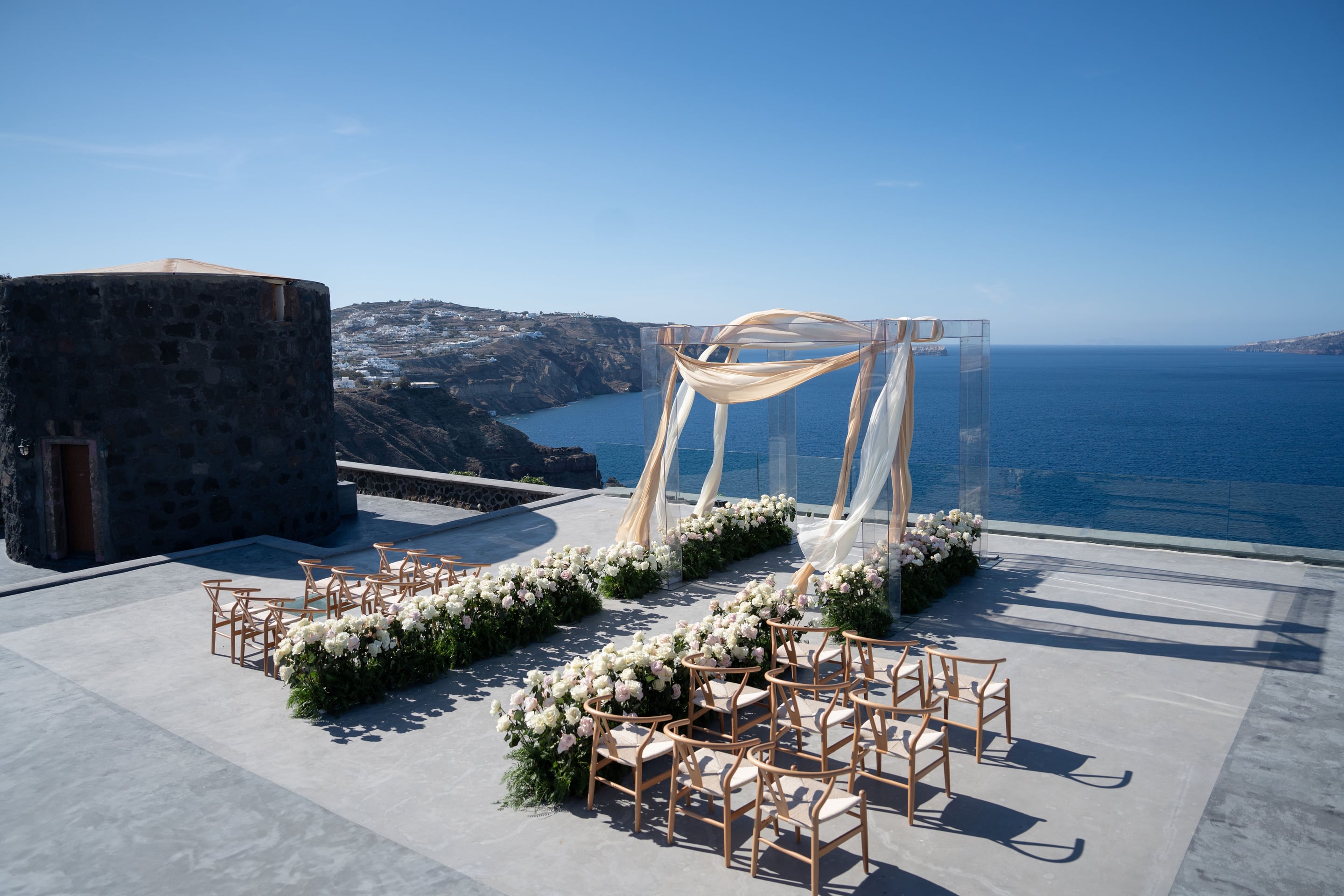 A wedding ceremony set up on a clifftop terrace at Cavo Ventus in Santorini, Greece, with two long aisles of white and blush roses and greenery leading to a clear acrylic arch draped in cream and champagne chiffon. Wooden wishbone chairs line the aisles, and beyond the terrace the deep blue Aegean Sea stretches to the horizon under a clear sky, with white-washed village houses clinging to the caldera cliffs on the left.