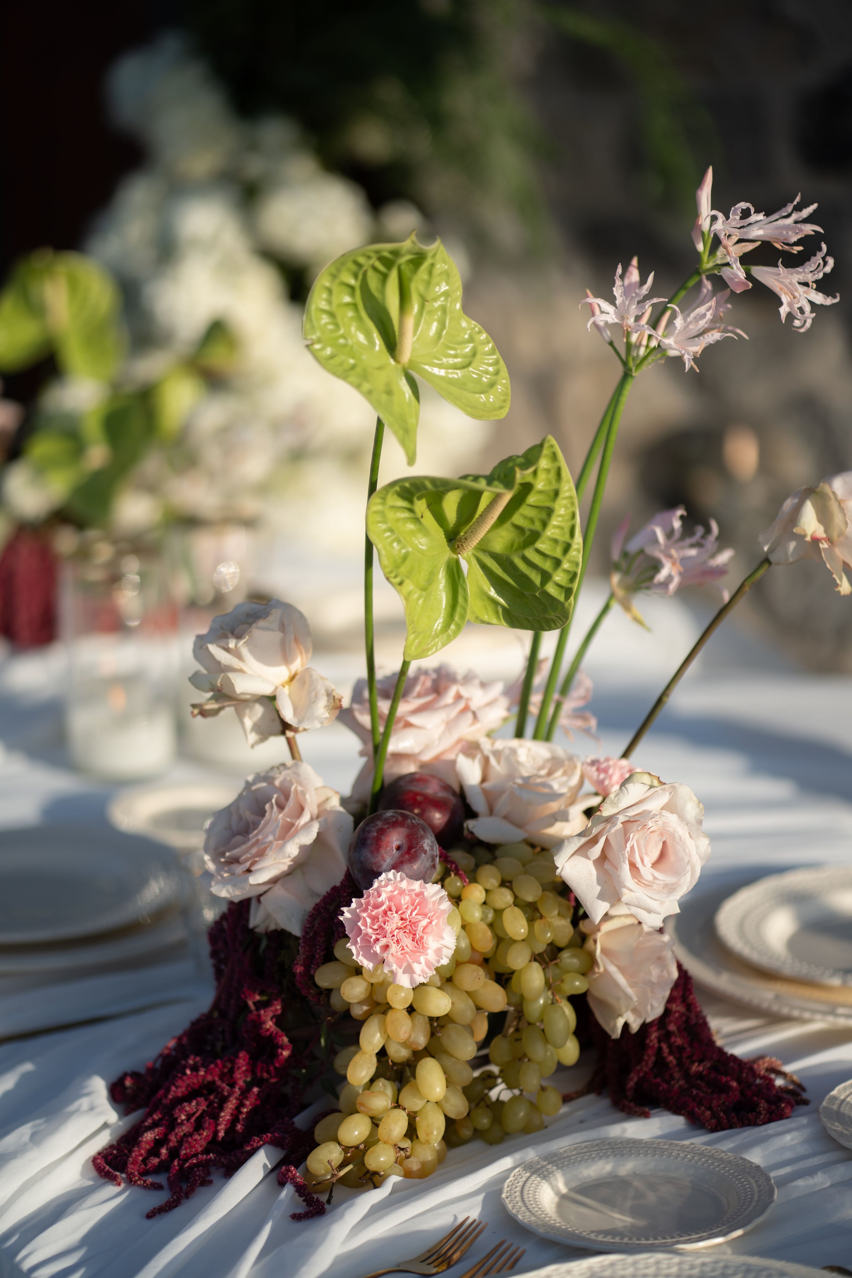 A wedding table centrepiece on a white linen cloth at Cavo Ventus in Greece, built around pale blush garden roses, a pink carnation and a tall green anthurium leaf. Clusters of pale green grapes and two dark plums spill from the base, trailed with deep burgundy amaranthus, and the soft golden light and blurred plates suggest a relaxed outdoor dinner.