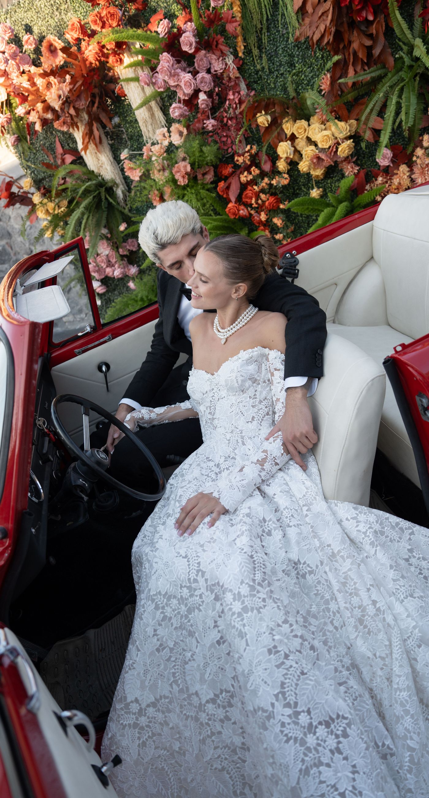 Isabella & Oliver - Under a clear blue sky, what unfolds is an ethereal expression of elegance and romance. The serene backdrop of vast, azure waters beautifully contrasts with the soft, delicate whites of the cascading petals, creating a scene both harmonious and enchanting. Meticulously arranged roses in gentle pinks and creams line the path, complementing the timeless lace and flowing structure of the bride's gown, while the groom's classic black suit adds a touch of refined sophistication. The graceful drapery, kissed by the gentle breeze, frames this moment of joy and forms a masterpiece of floral artistry, balancing nature's serenity with bridal grandeur. - Photo 6