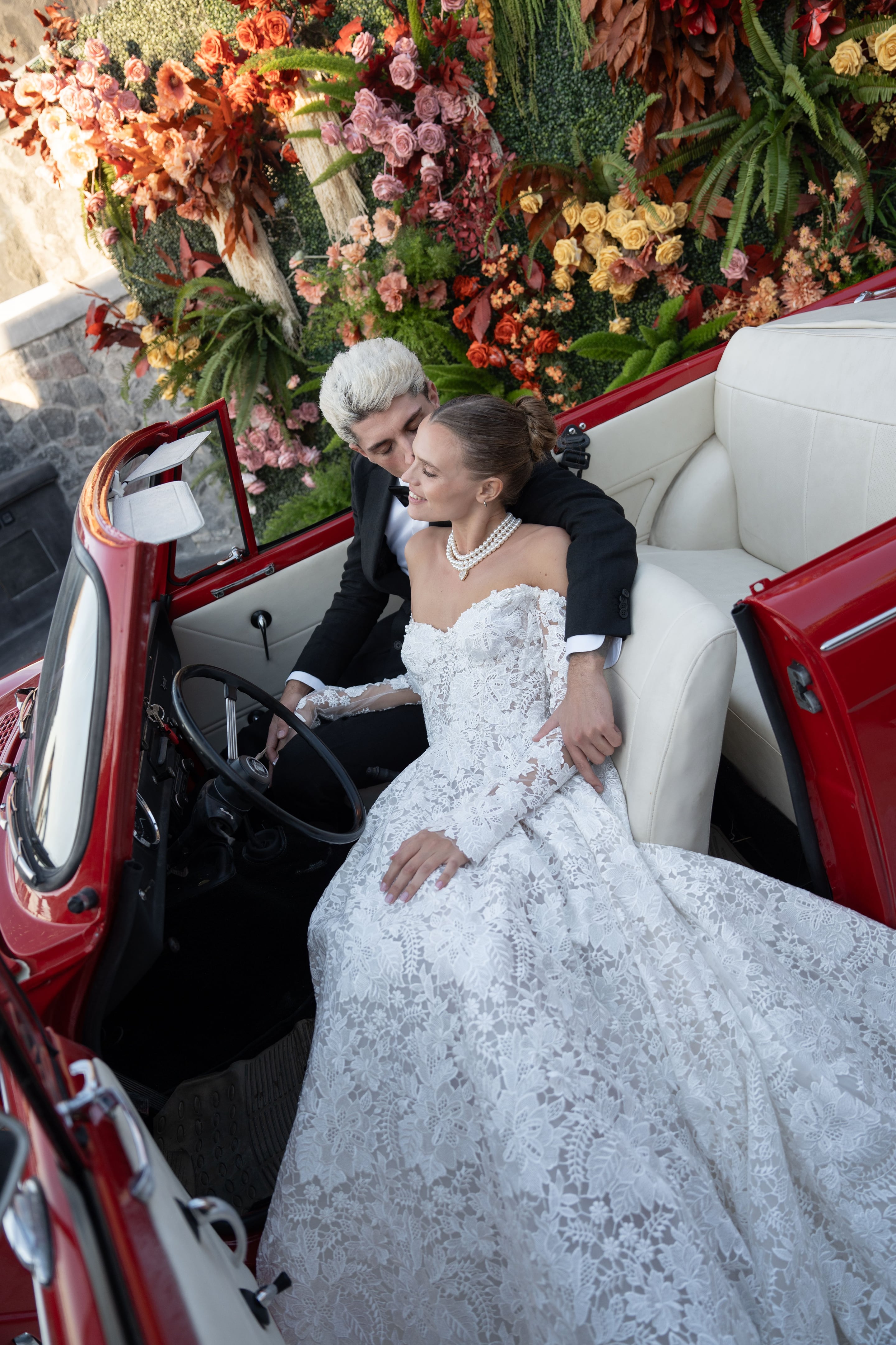 A bride in a white lace off-the-shoulder gown and a multi-strand pearl necklace sits smiling in the cream leather seat of a vintage red convertible, while a man with silver-white hair in a black suit leans in behind her, his eyes closed. Behind them is a wall of wedding flowers in terracotta, peach and blush roses, dried russet foliage and golden yellow roses, set at Cavo Ventus in Greece.