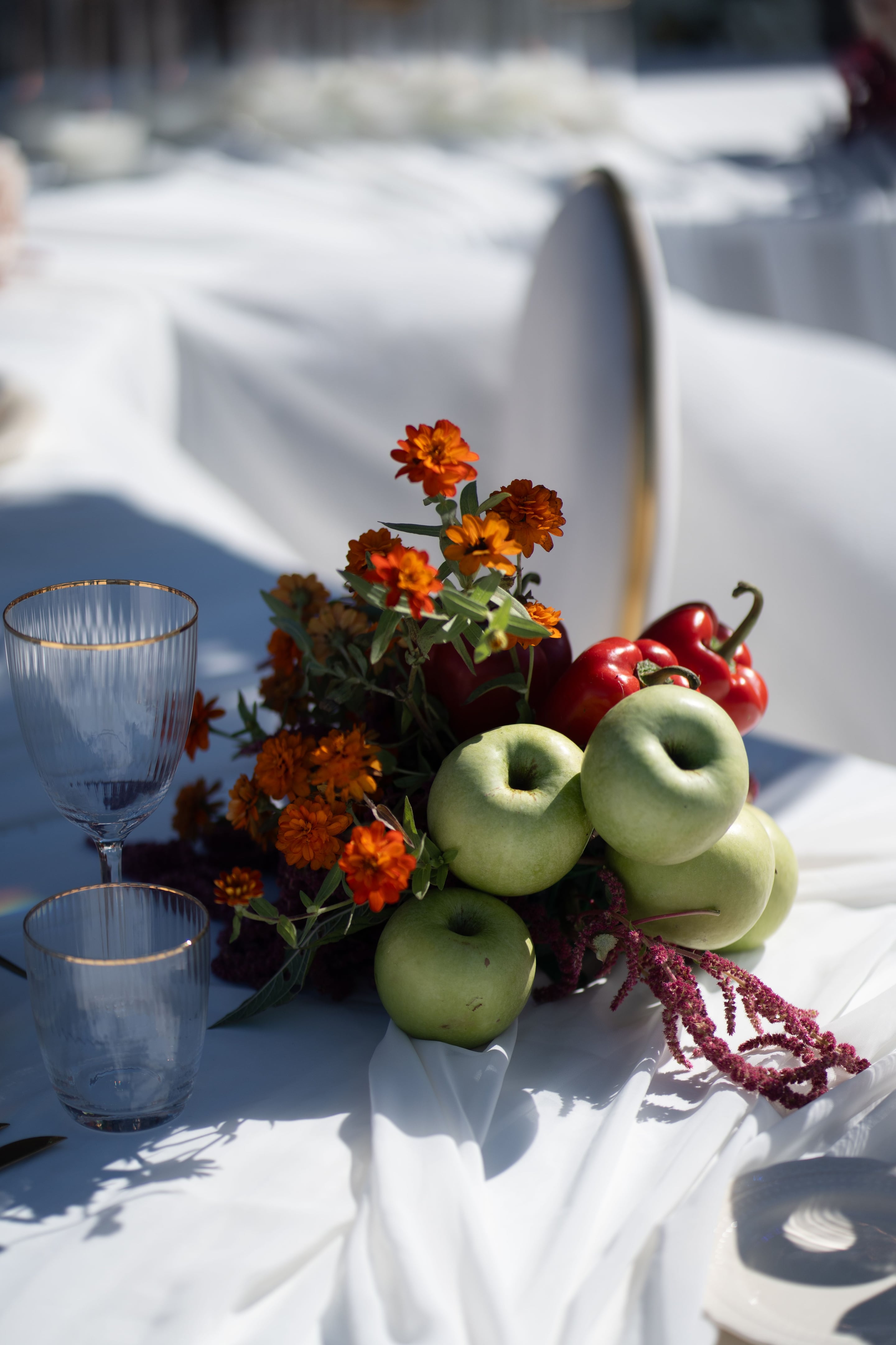 A wedding table centrepiece at Cavo Ventus in Greece, built around a cluster of green apples and glossy red peppers heaped beside a loose bunch of orange zinnias, dark burgundy amaranthus trailing down, and slender olive-toned foliage. Two ribbed crystal wine glasses with gold rims stand at the left edge on a white linen cloth, with warm sunlight casting soft shadows across the table. The mood is rustic, autumnal and relaxed.