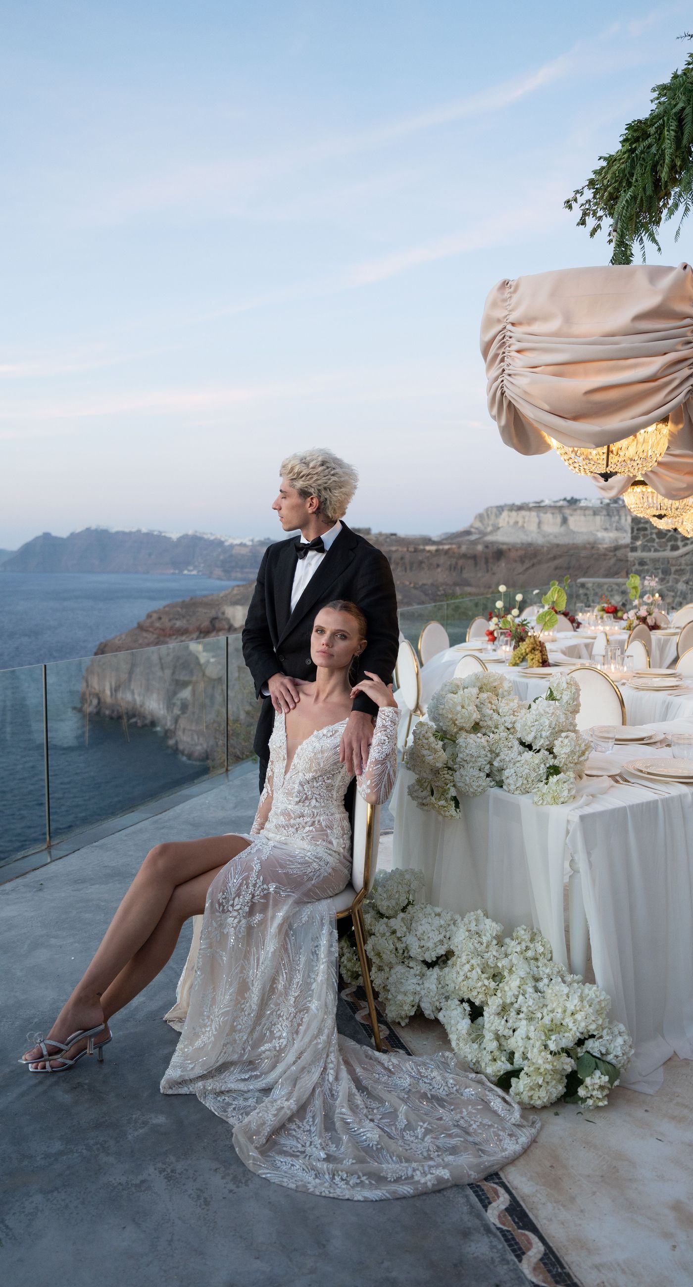 Isabella & Oliver - Under a clear blue sky, what unfolds is an ethereal expression of elegance and romance. The serene backdrop of vast, azure waters beautifully contrasts with the soft, delicate whites of the cascading petals, creating a scene both harmonious and enchanting. Meticulously arranged roses in gentle pinks and creams line the path, complementing the timeless lace and flowing structure of the bride's gown, while the groom's classic black suit adds a touch of refined sophistication. The graceful drapery, kissed by the gentle breeze, frames this moment of joy and forms a masterpiece of floral artistry, balancing nature's serenity with bridal grandeur. - Photo 17