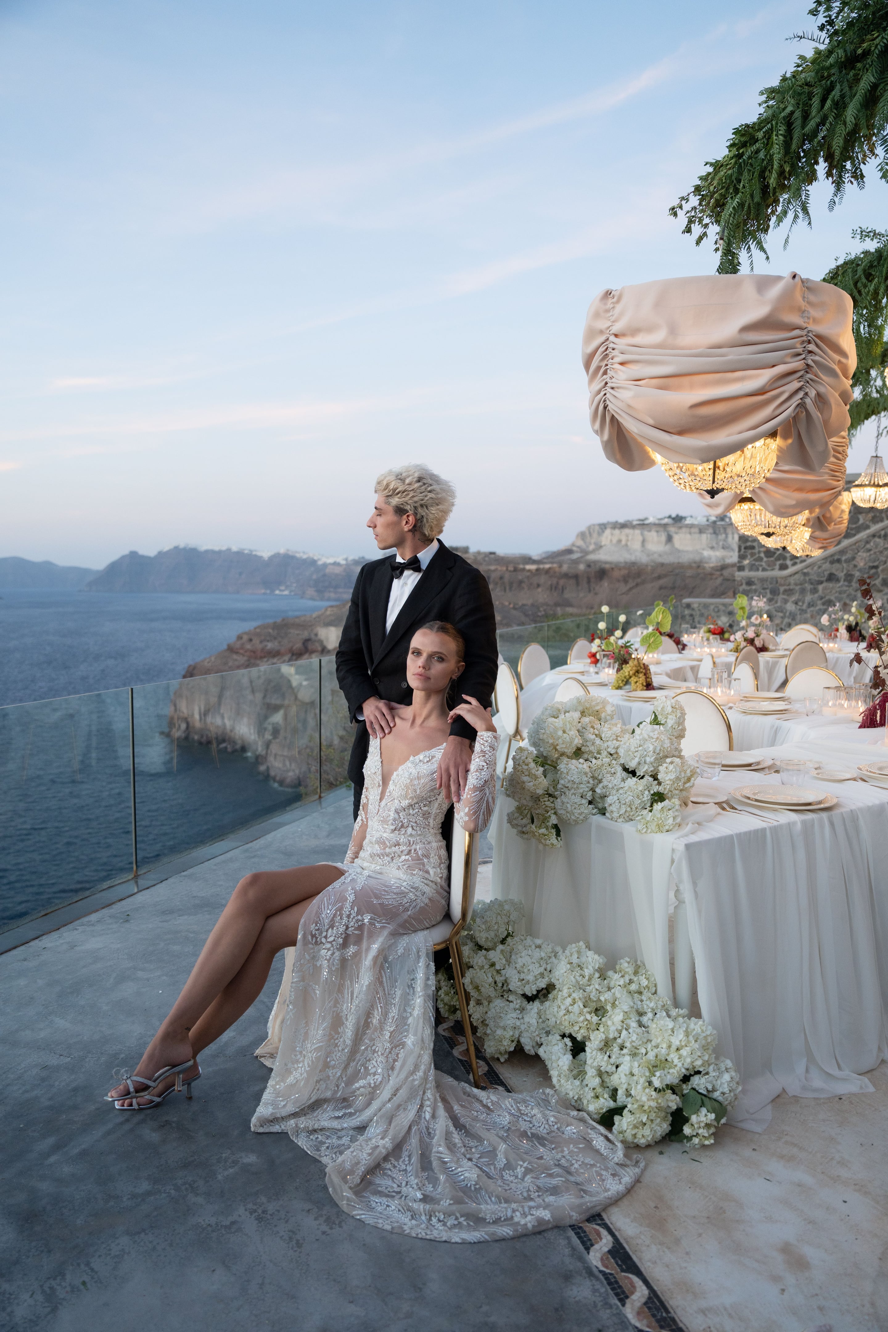 A bride in a sheer, lace-embroidered gown sits on a gold-framed chair at the edge of a clifftop terrace, while a groom in a black tuxedo and bow tie stands behind her with a hand on her shoulder, both gazing out over a calm blue sea at dusk. Behind them, a long white-draped banquet table is set with white hydrangeas and candlelight, under gathered beige fabric and crystal chandeliers, at Cavo Ventus on Santorini, Greece.
