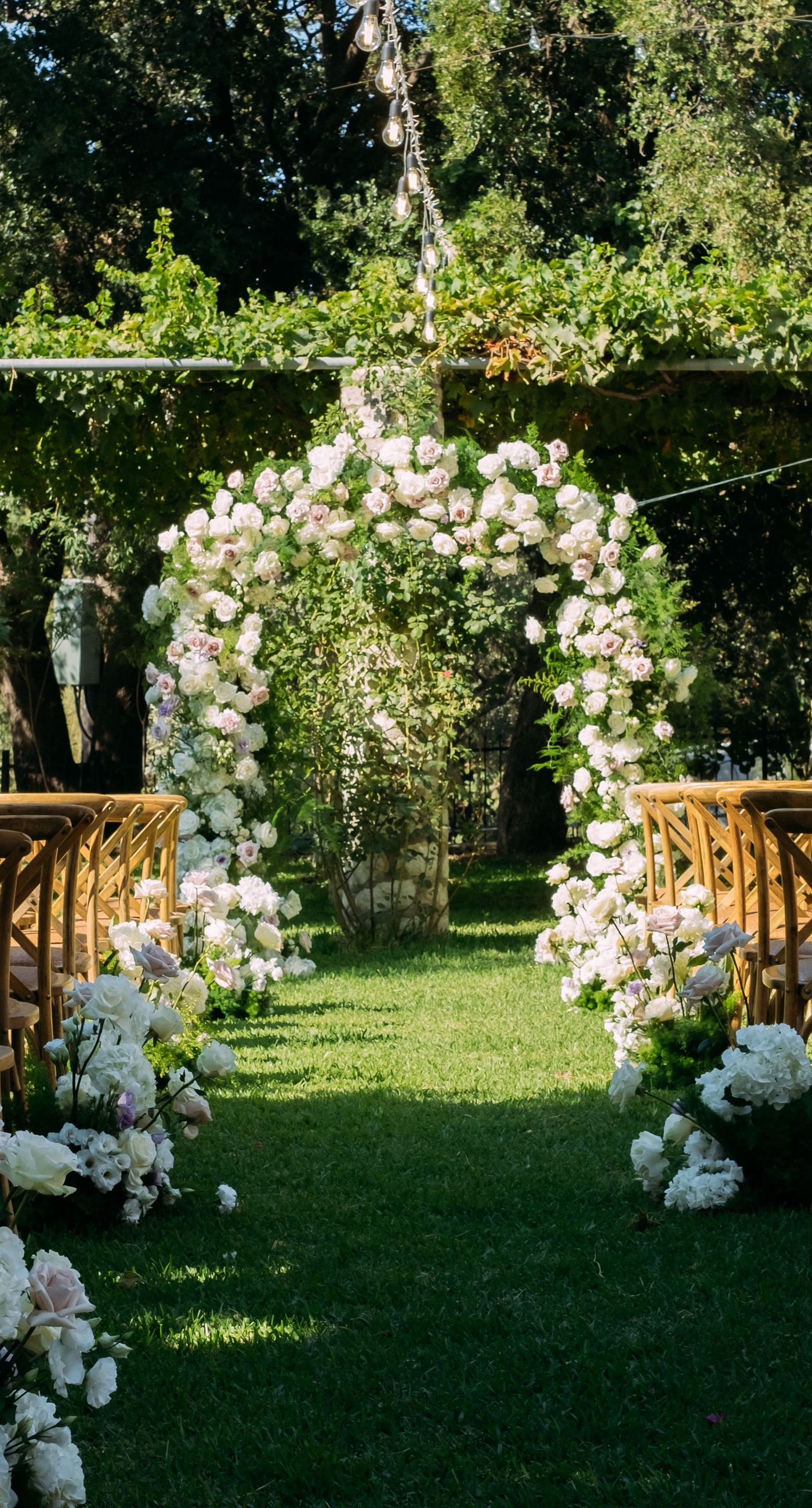 June & Tim - Beneath a lush canopy of greenery, a symphony of ivory and blush blooms cascades in a breathtaking floral arch. The delicate roses and verdant ferns intertwine effortlessly, creating a harmonious backdrop that exudes elegance and romance. The serene garden setting bathes the scene in soft, natural light, enhancing the ethereal beauty of the arrangement. The joyful expressions of the couple, captured in this radiant moment, complement the stunning florals with a vibrant energy. This captivating work reflects a masterful blend of nature's artistry and human emotion, making it an unforgettable statement in any portfolio. - Photo 1
