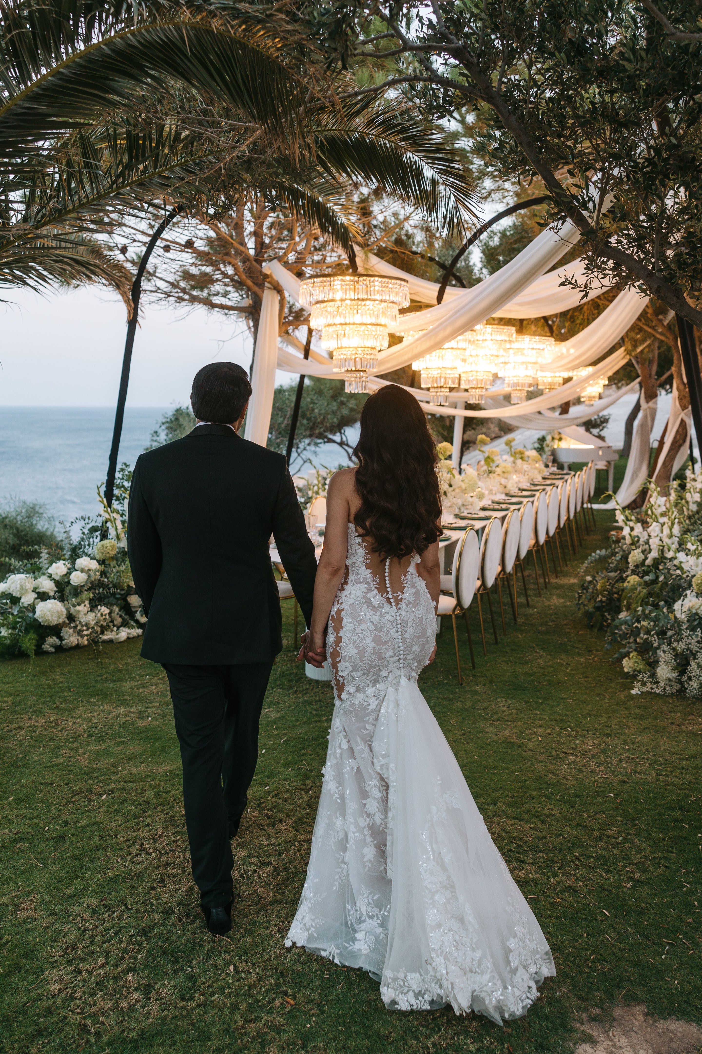 A bride with long wavy dark hair and a lace-covered backless gown walks hand in hand with a groom in a dark suit, seen from behind, along a clifftop lawn at Cycladic Gem in Greece. Above them hang tiered crystal chandeliers under draped white fabric, and a long banquet table is lined with white flowers, white hydrangeas and cream blooms, with the Aegean Sea glowing blue beyond the olive and palm trees.
