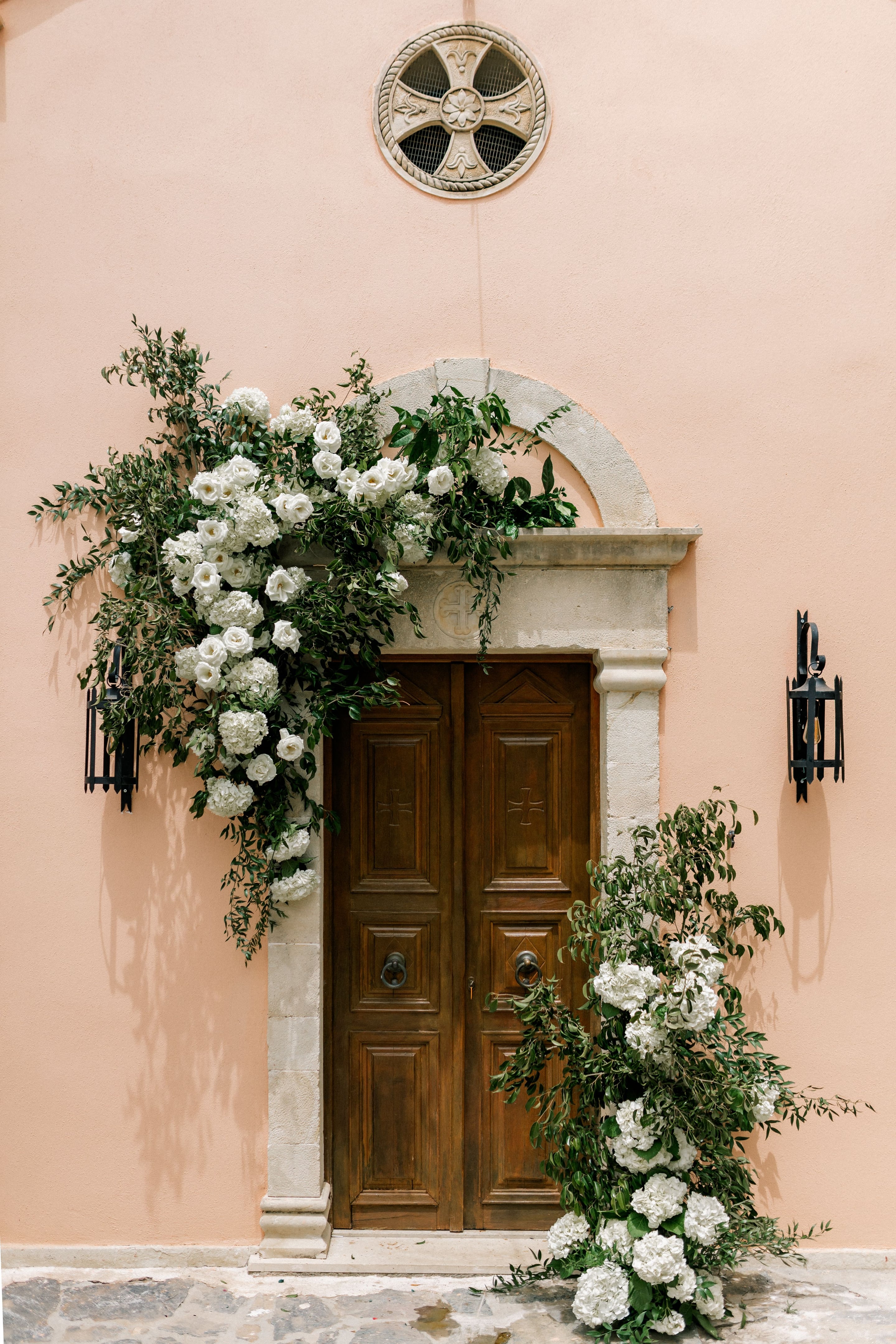 A pair of tall wooden double doors with carved panels and iron ring handles, set in a pale stone arch against a peach-coloured wall, is framed by lush cascading flowers. White lisianthus, white hydrangeas and small roses spill down the left side of the arch in a loose, trailing curve, woven through green olive-like foliage, while a second garland of white hydrangeas and greenery hangs beside the right door. This is wedding floral design at Agreco Farms in Greece.