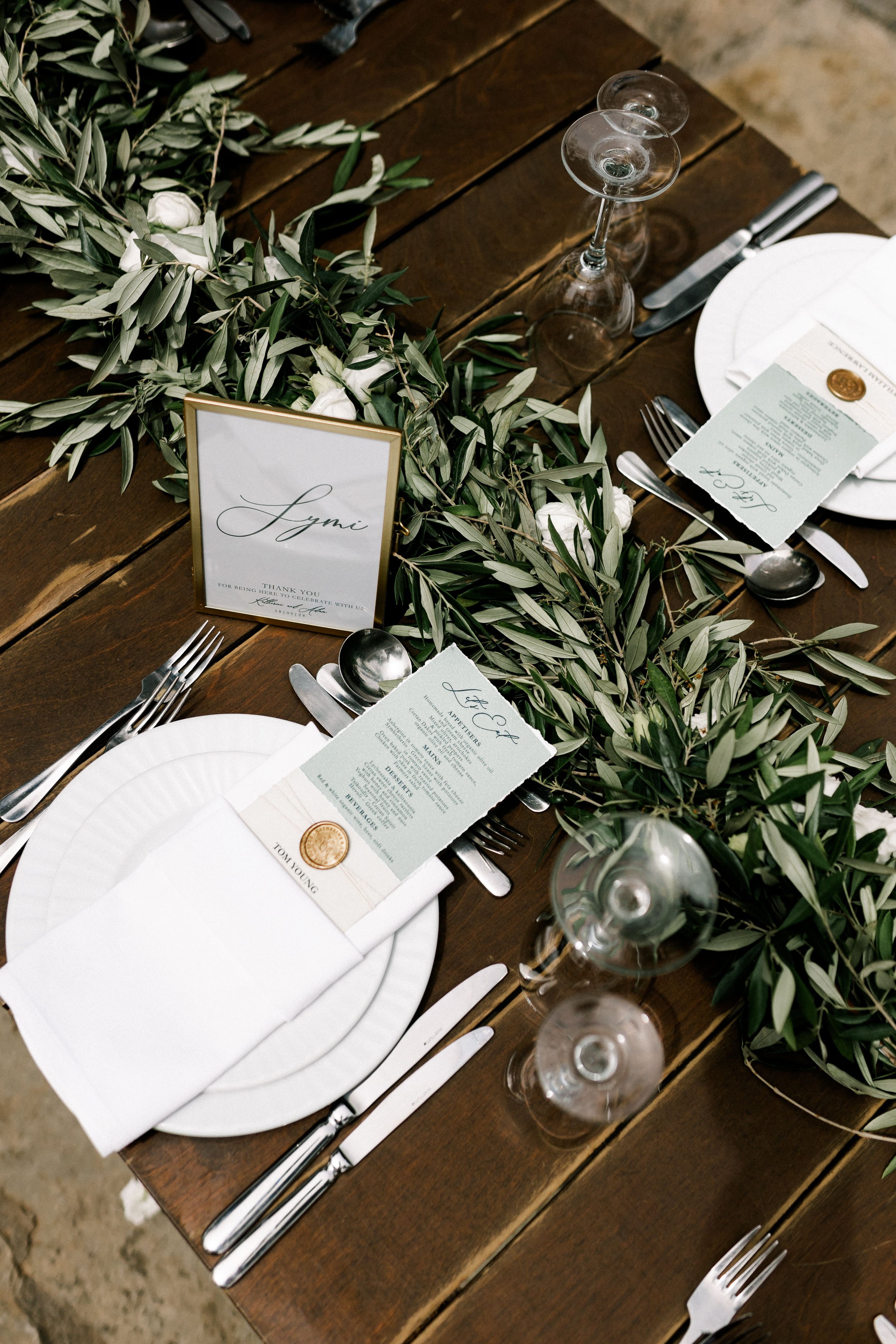 A wooden banquet table at Agreco Farms in Greece is dressed with a long runner of silvery olive branches studded with small white garden roses, set for a wedding meal. In the foreground a white plate holds a folded linen napkin and a menu card in sage green, sealed with a gold wax stamp and bearing a guest's name. A framed gold place card reading "Lymi" sits among the greenery, with silver cutlery and stemmed glasses catching the soft natural light.