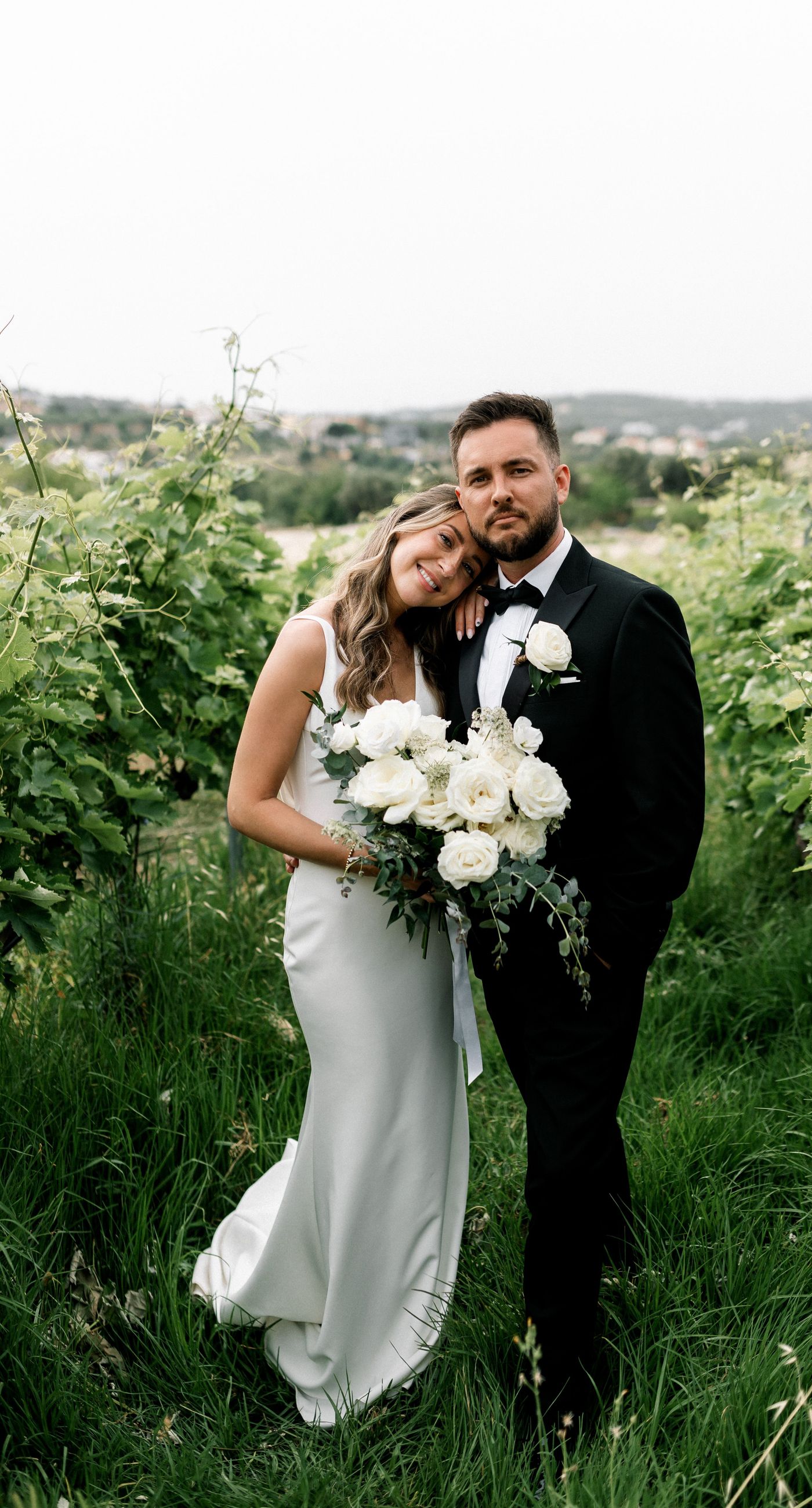 Katherine & Adam - Nestled within the lush, sprawling rows of a vineyard, the scene captures an exquisite moment of romance and elegance. The verdant landscape creates a serene backdrop, perfectly complementing the delicate whites and soft greens of the bride's bouquet. The interplay between the timeless black tuxedo and the pure white gown establishes a harmonious balance, inviting you into a world of classic beauty and serene intimacy. The gentle kiss shared amidst the verdant vines evokes a sense of rustic charm blended with refined grace, an ideal setting for a florist's artistry in capturing life's tender moments. - Photo 15
