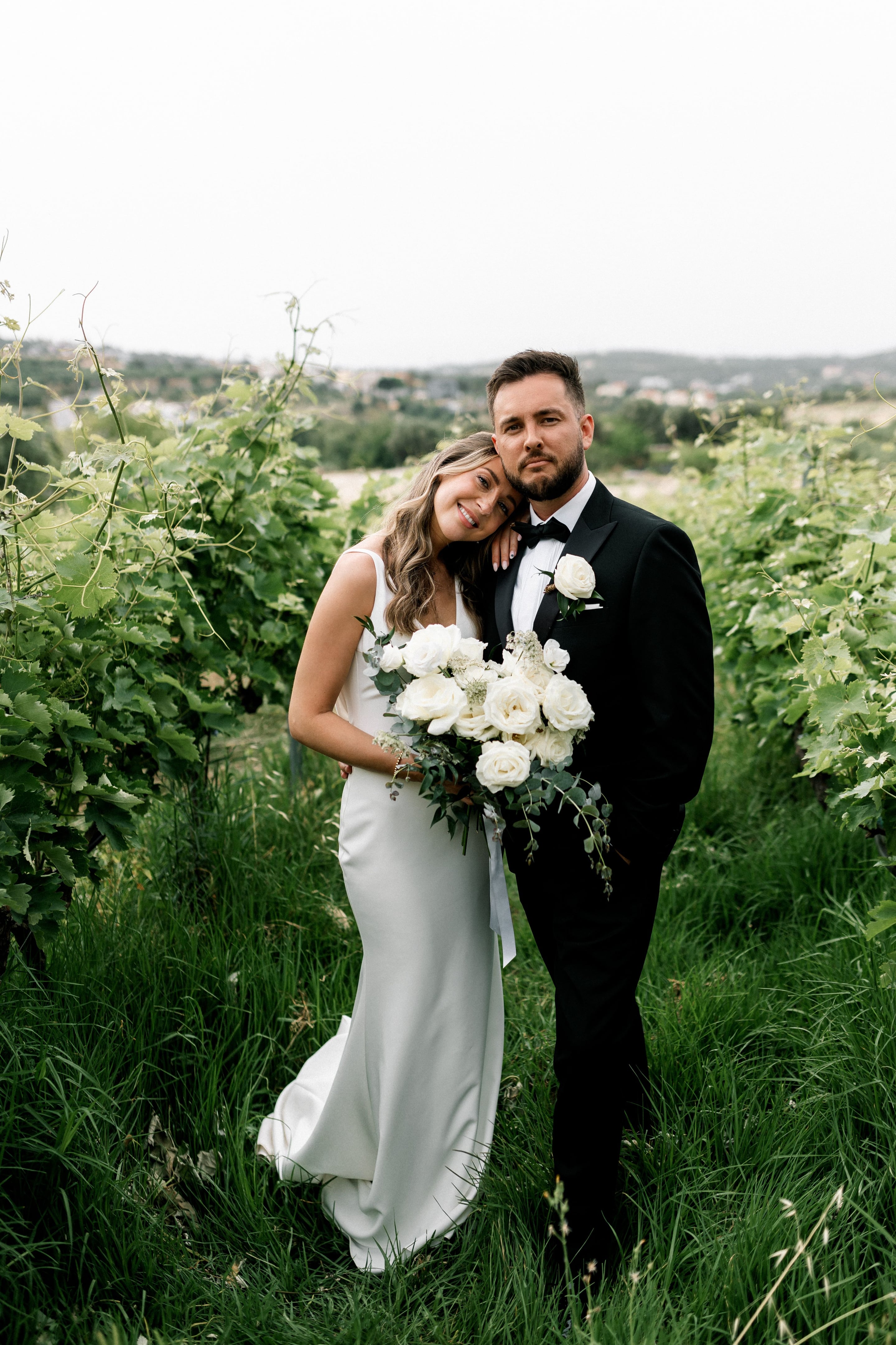 A bride in a simple satin column gown leans her head affectionately against her groom, who wears a black tuxedo with a bow tie and a white rose boutonniere. She holds a loose bouquet of white garden roses, ranunculus and airy white blooms with trailing eucalyptus, tied with a long ivory ribbon. They stand among green grapevines and tall grass at Agreco Farms, with soft hills and houses blurred behind them, a wedding bouquet and groom's flowers in Greece.