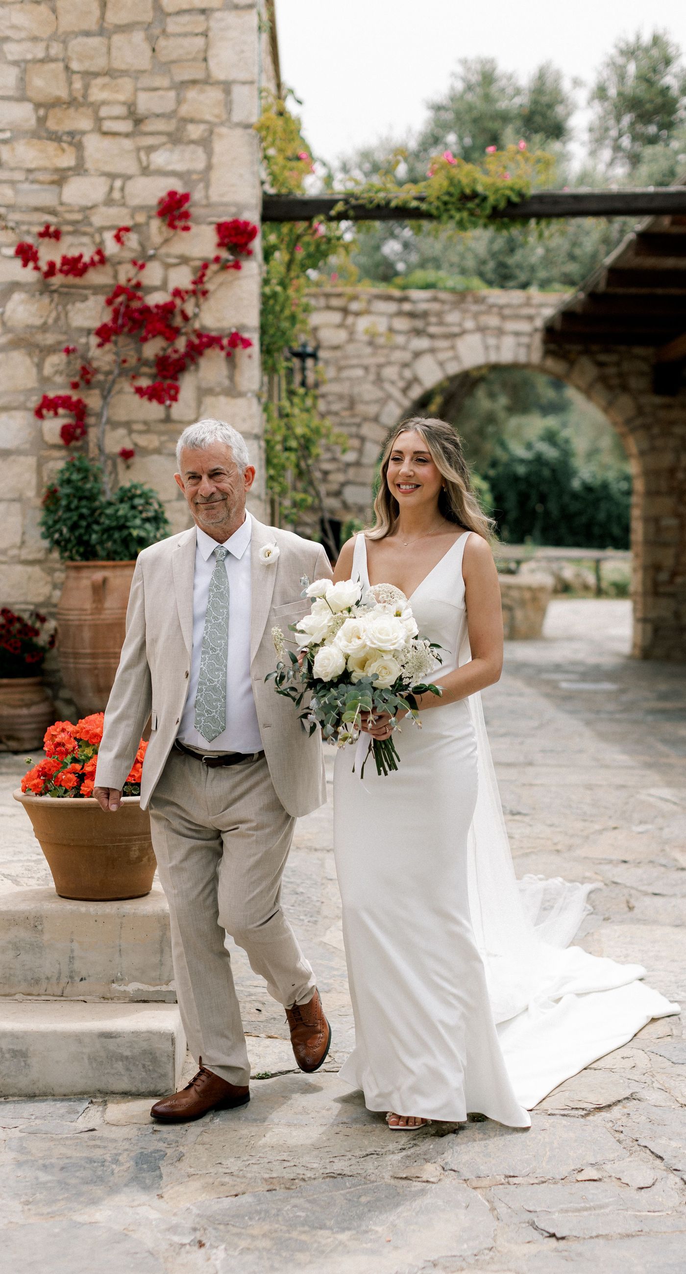 Katherine & Adam - Nestled within the lush, sprawling rows of a vineyard, the scene captures an exquisite moment of romance and elegance. The verdant landscape creates a serene backdrop, perfectly complementing the delicate whites and soft greens of the bride's bouquet. The interplay between the timeless black tuxedo and the pure white gown establishes a harmonious balance, inviting you into a world of classic beauty and serene intimacy. The gentle kiss shared amidst the verdant vines evokes a sense of rustic charm blended with refined grace, an ideal setting for a florist's artistry in capturing life's tender moments. - Photo 4