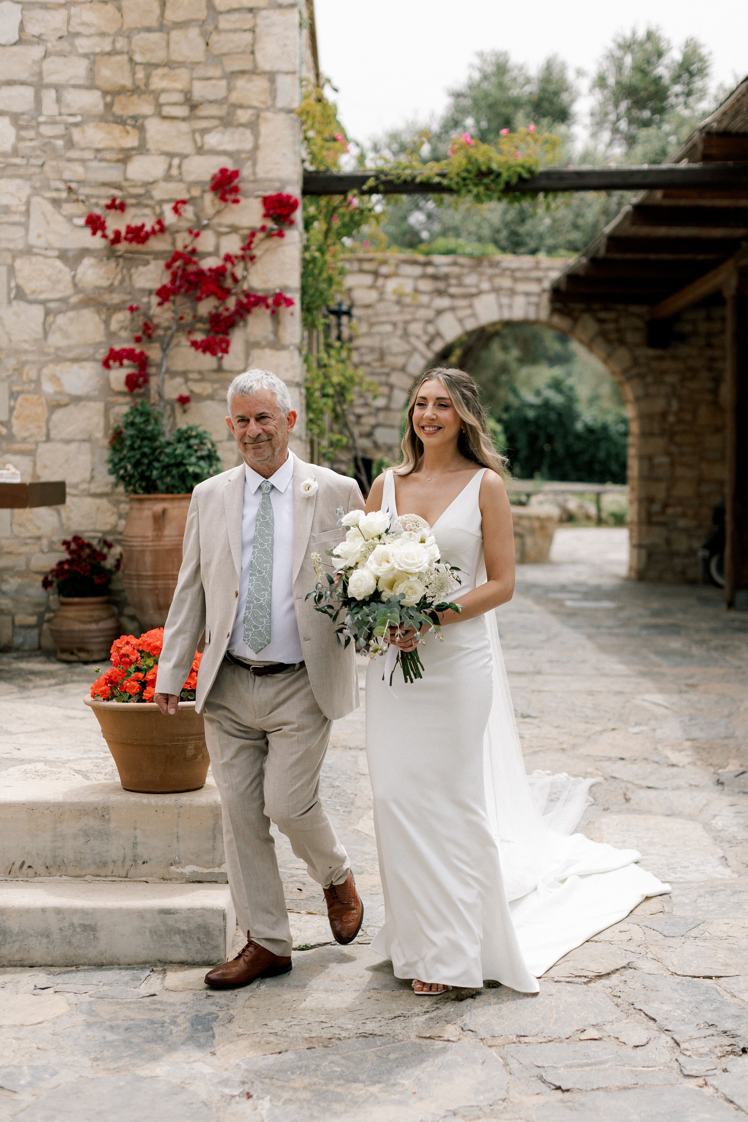 A bride in a sleek white crepe gown with a deep V-neck walks beside an older man in a pale stone-coloured suit and sage tie along a flagstone courtyard at Agreco Farms in Greece. She holds a loose bridal bouquet of white garden roses, white ranunculus and feathery queen anne's lace, edged with silver-green eucalyptus. Behind them, a rounded stone archway, red bougainvillea climbing the rough stone wall and terracotta pots of orange blooms create a warm, sunlit wedding scene.