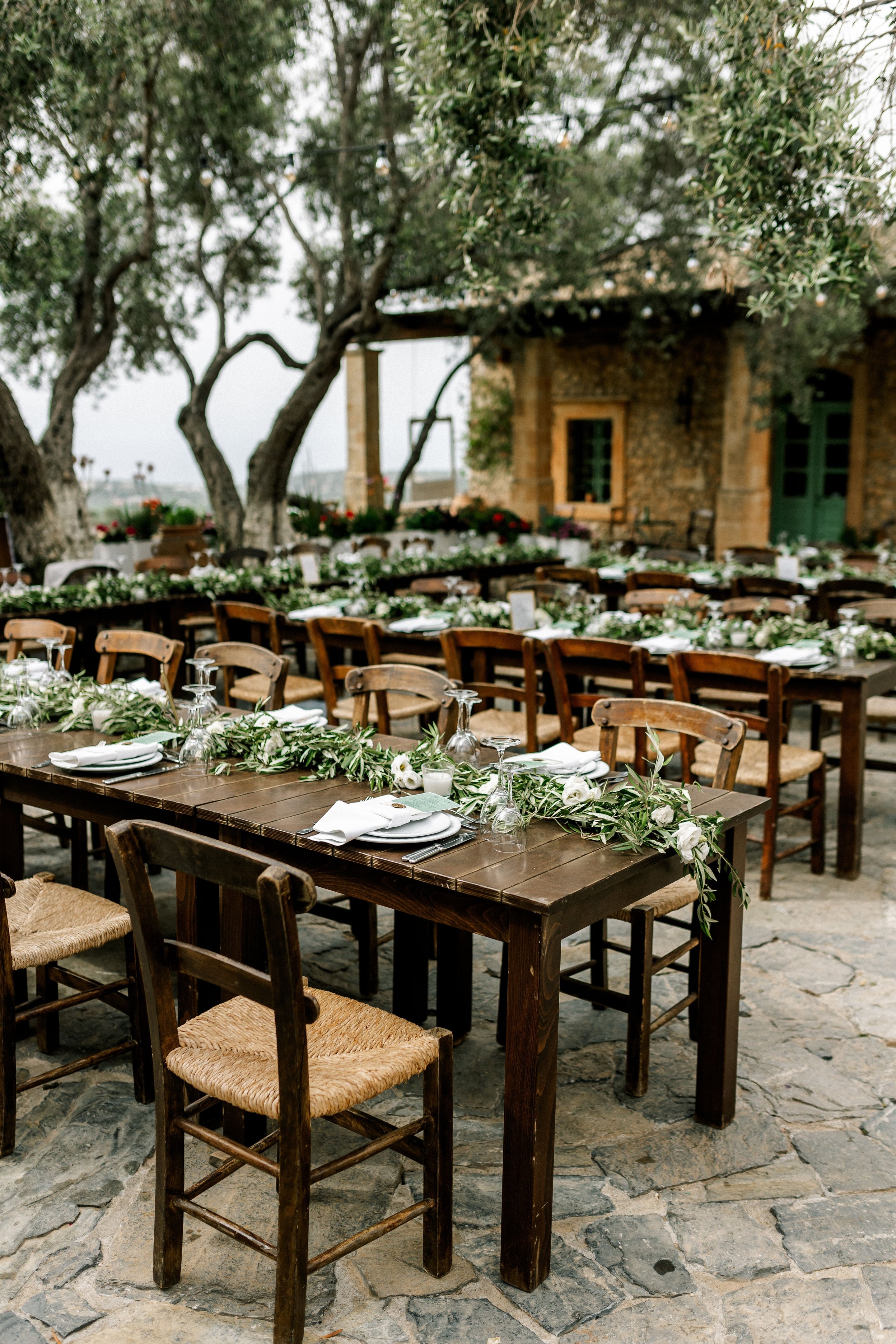 Long rustic wooden tables on a stone terrace under ancient olive trees are dressed with runners of silvery olive foliage and white garden roses, with white napkins, plates and inverted wine glasses laid out for a wedding meal at Agreco Farms in Greece. Strings of festoon lights hang above, and a honey-coloured stone farmhouse with a green door frames the scene, while the soft grey sky gives the whole setting a calm, natural mood.