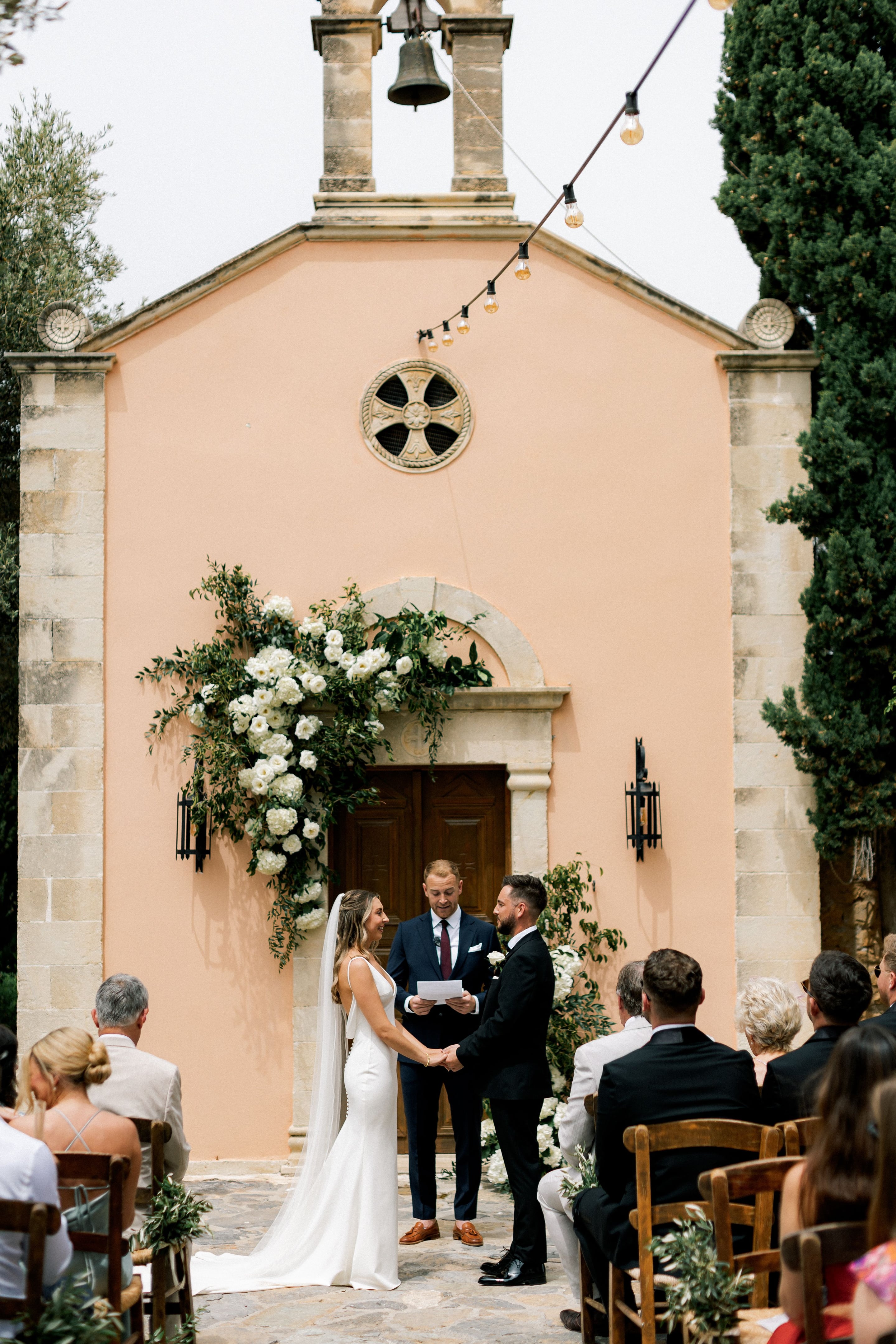 A wedding ceremony in Greece at Agreco Farms, where a bride in a fitted white gown with a long veil and a groom in a black suit hold hands facing each other beneath a cascading arch of white roses, hydrangeas and trailing green foliage. The arch frames a wooden double door set in a peach-coloured wall with a stone surround. Guests sit on rustic wooden chairs in the warm, bright courtyard, decorated with olive sprigs along the aisle.