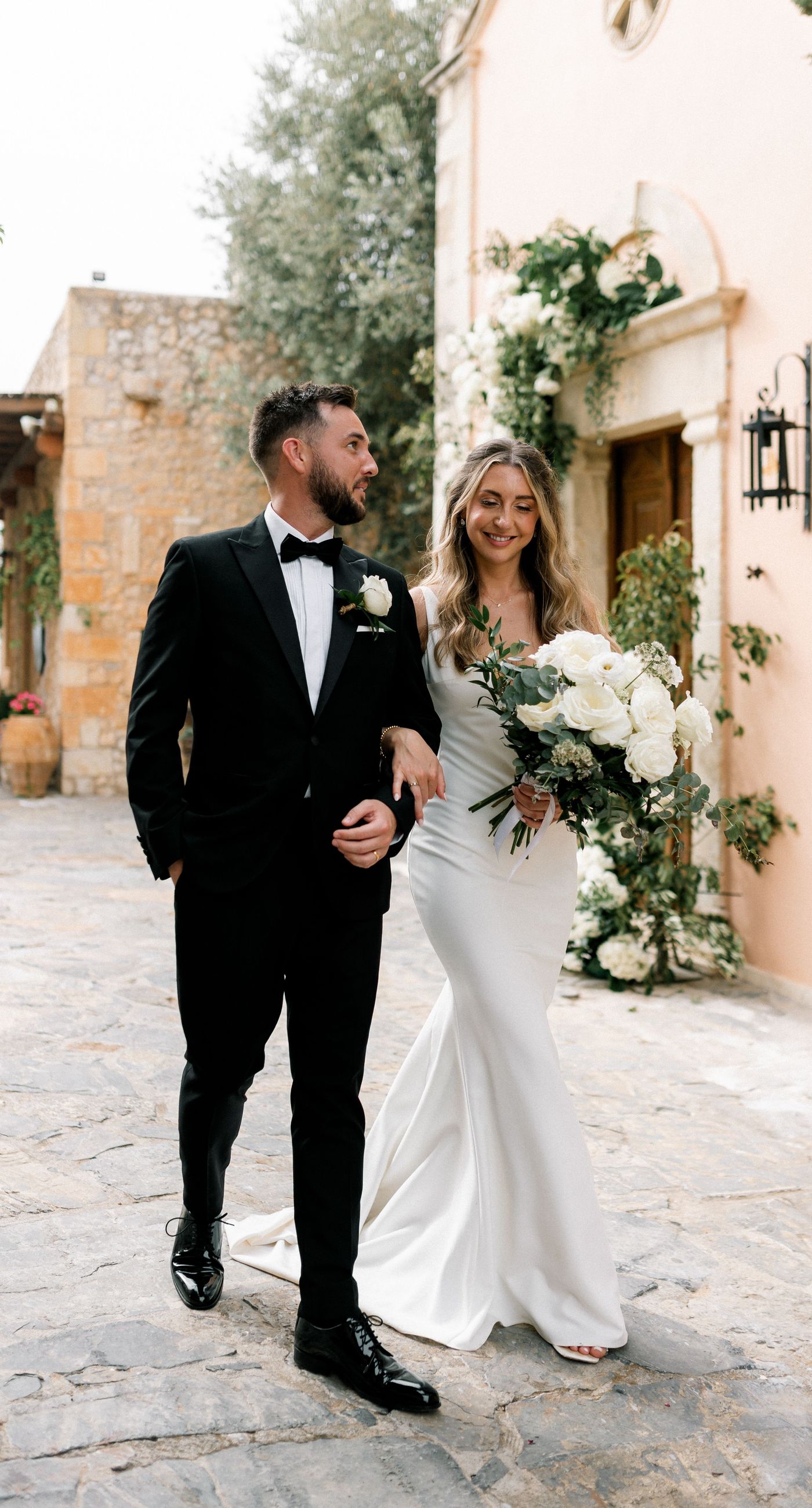 Katherine & Adam - Nestled within the lush, sprawling rows of a vineyard, the scene captures an exquisite moment of romance and elegance. The verdant landscape creates a serene backdrop, perfectly complementing the delicate whites and soft greens of the bride's bouquet. The interplay between the timeless black tuxedo and the pure white gown establishes a harmonious balance, inviting you into a world of classic beauty and serene intimacy. The gentle kiss shared amidst the verdant vines evokes a sense of rustic charm blended with refined grace, an ideal setting for a florist's artistry in capturing life's tender moments. - Photo 9
