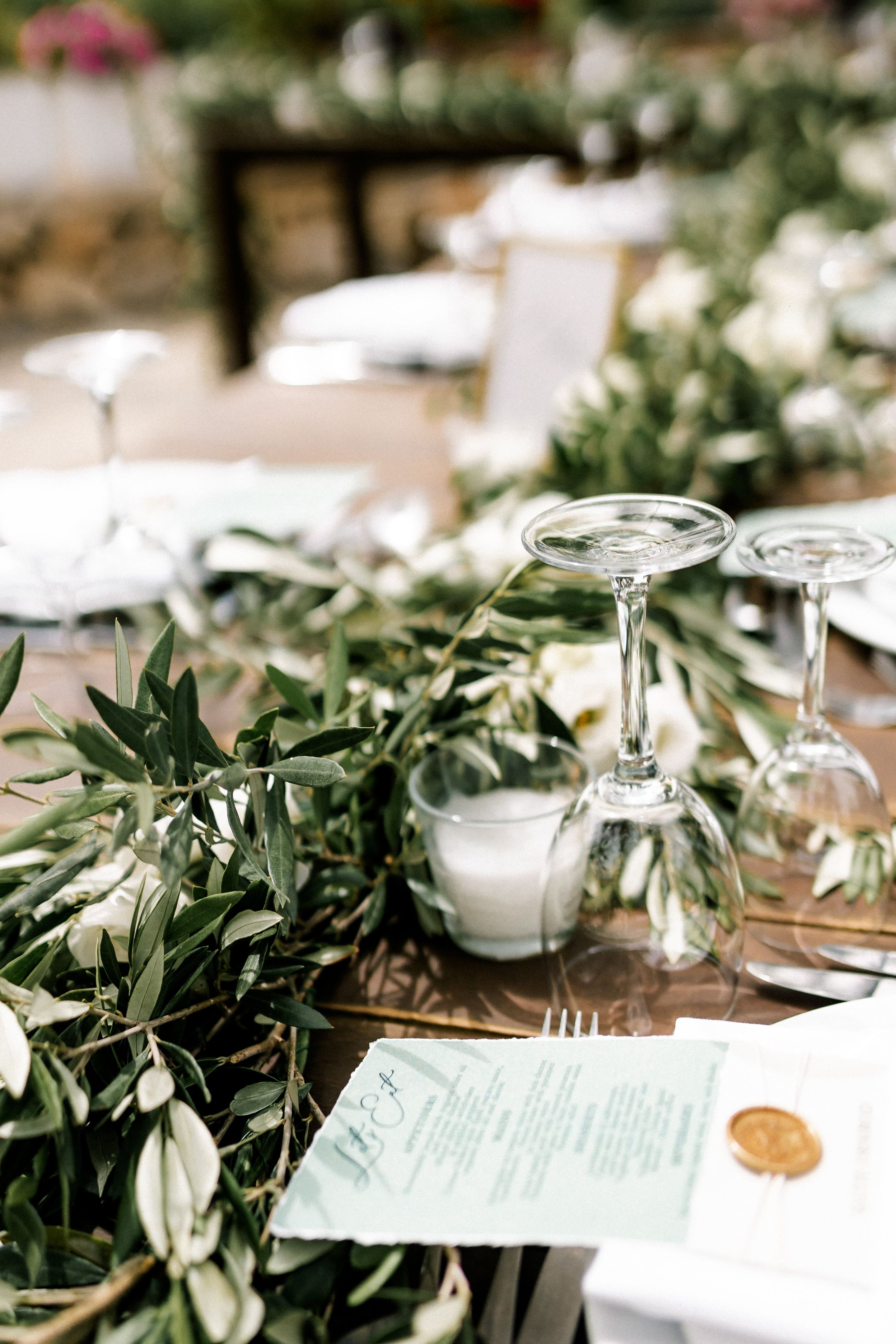 Upturned wine glasses sit on a wooden wedding table at Agreco Farms in Greece, framed by a long runner of olive branches and white blooms that look like garden roses. Beside them is a small glass votive candle, and a pale green menu card rests on a white napkin near the cutlery. The soft, out-of-focus rows of tables stretching behind suggest a bright, airy reception.