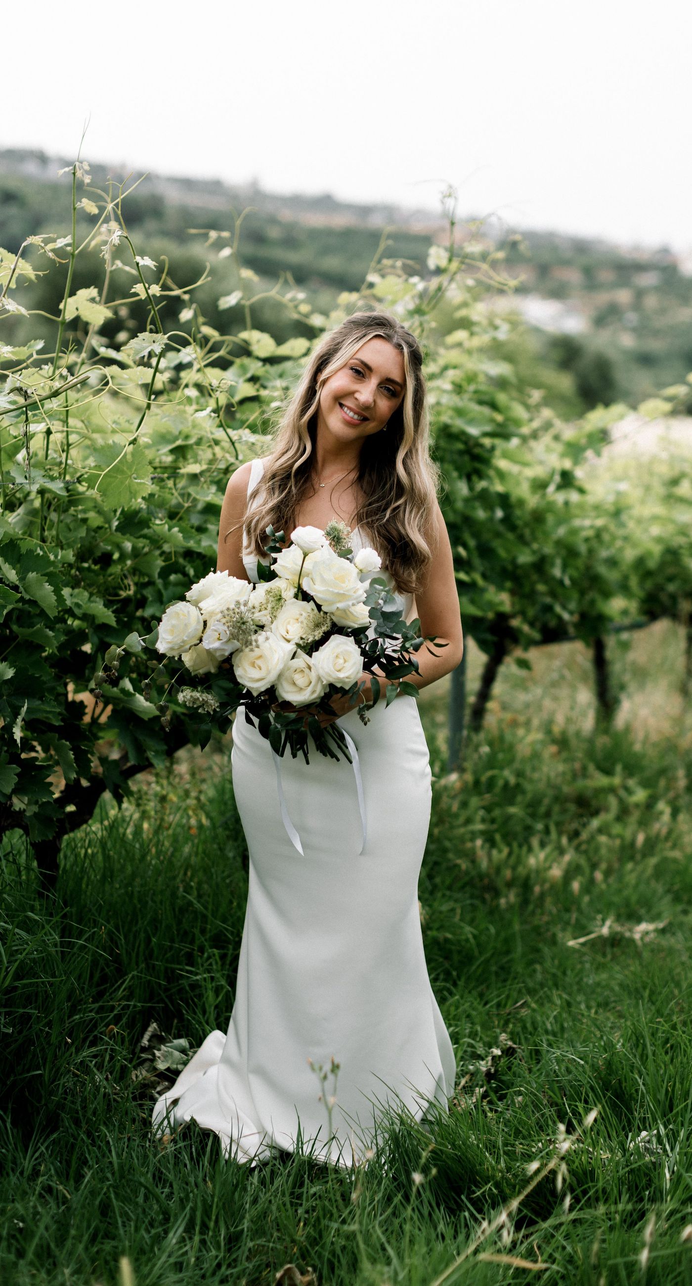 Katherine & Adam - Nestled within the lush, sprawling rows of a vineyard, the scene captures an exquisite moment of romance and elegance. The verdant landscape creates a serene backdrop, perfectly complementing the delicate whites and soft greens of the bride's bouquet. The interplay between the timeless black tuxedo and the pure white gown establishes a harmonious balance, inviting you into a world of classic beauty and serene intimacy. The gentle kiss shared amidst the verdant vines evokes a sense of rustic charm blended with refined grace, an ideal setting for a florist's artistry in capturing life's tender moments. - Photo 14
