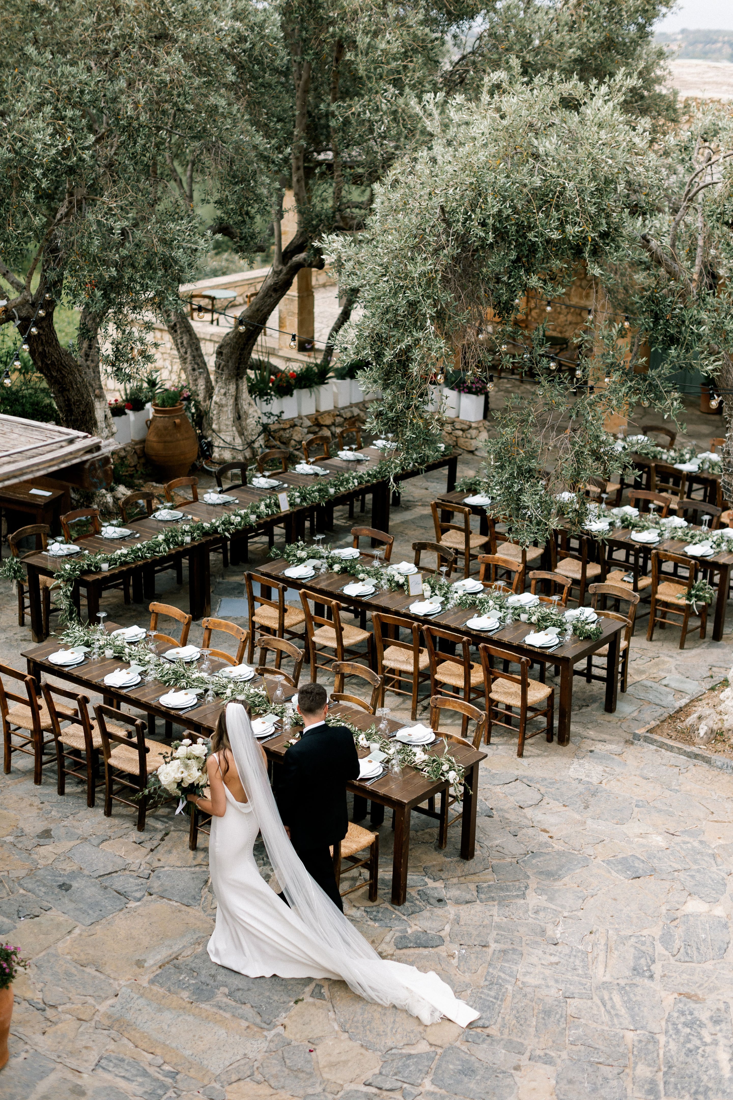 A high aerial view of a stone-paved courtyard at Agreco Farms in Greece, where long rustic wooden banquet tables run in rows beneath gnarled olive trees strung with warm festoon lights. Each table is dressed with trailing olive branches, white blooms and folded white napkins, with rush-seat chairs drawn up around them. In the foreground a bride in a fitted white gown and long veil, holding a white rose bouquet, walks beside her groom in a dark suit toward the tables, as wedding guests and staff prepare for the reception.