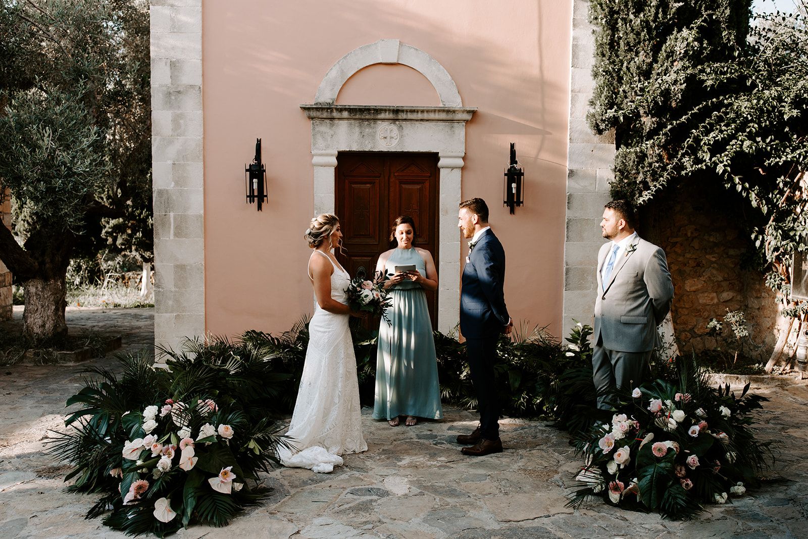 Two palms-and-blush floral installations frame a small wedding ceremony in front of a peach-washed chapel with a carved stone doorway and a dark wooden door, in Greece. The bride in a lace gown holds a loose bouquet of pale pink and white blooms, while the groom in a navy suit faces her and a celebrant in a dusty-blue gown reads from a tablet. Warm late-afternoon light falls softly across the stone courtyard.