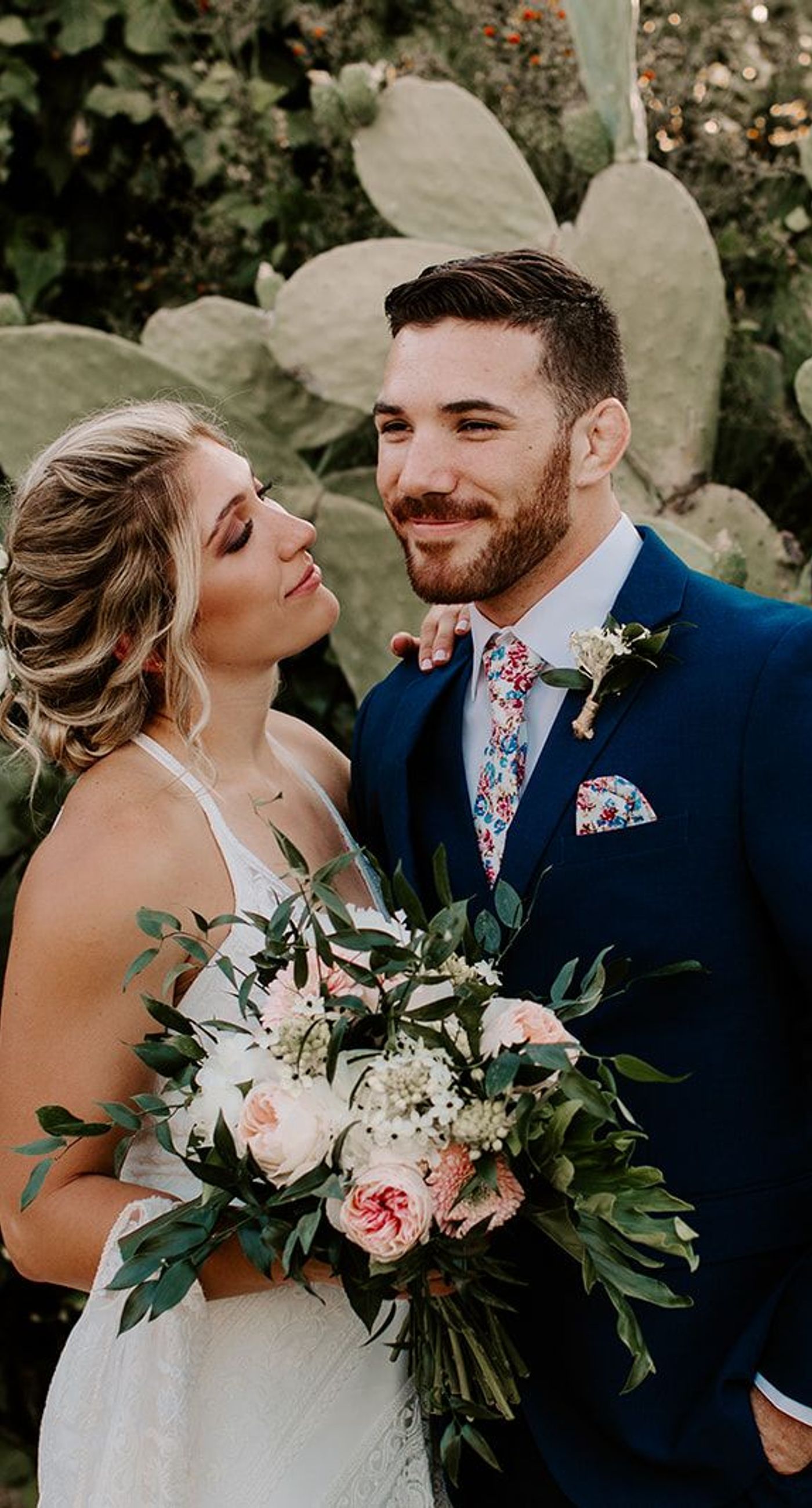 Ketty & Smith - In a symphony of soft pink hues and delicate floral arrangements, the romance of this work unfolds against a backdrop of timeless charm. The couple stands enveloped in a misty blush, their intimate embrace framed by an elegant stone archway, exuding elegance and warmth. The bride's bouquet, a harmonious blend of greenery and pastel blossoms, complements the sophisticated navy of the groom's suit, while the gentle palette of the setting evokes an ethereal, dreamlike quality. Every element is curated with graceful precision, creating a tapestry of love and beauty that is both refined and captivating. - Photo 2