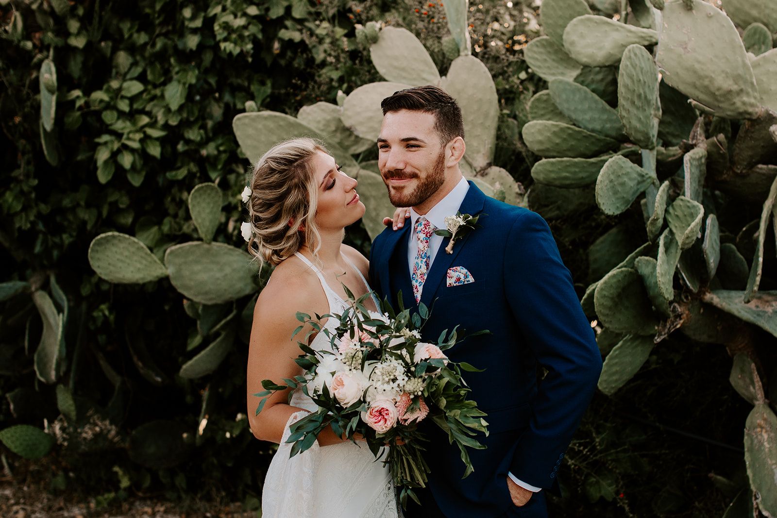 A bride with a soft updo and small white blooms in her hair leans her head toward a smiling groom in a navy suit with a floral tie and pocket square, standing before large pale-green prickly pear cactus pads. She holds a loose bridal bouquet of blush garden roses, white blossoms and trailing eucalyptus-style greenery. These are wedding flowers in Greece, and the warm, golden light makes the moment feel gentle and intimate.