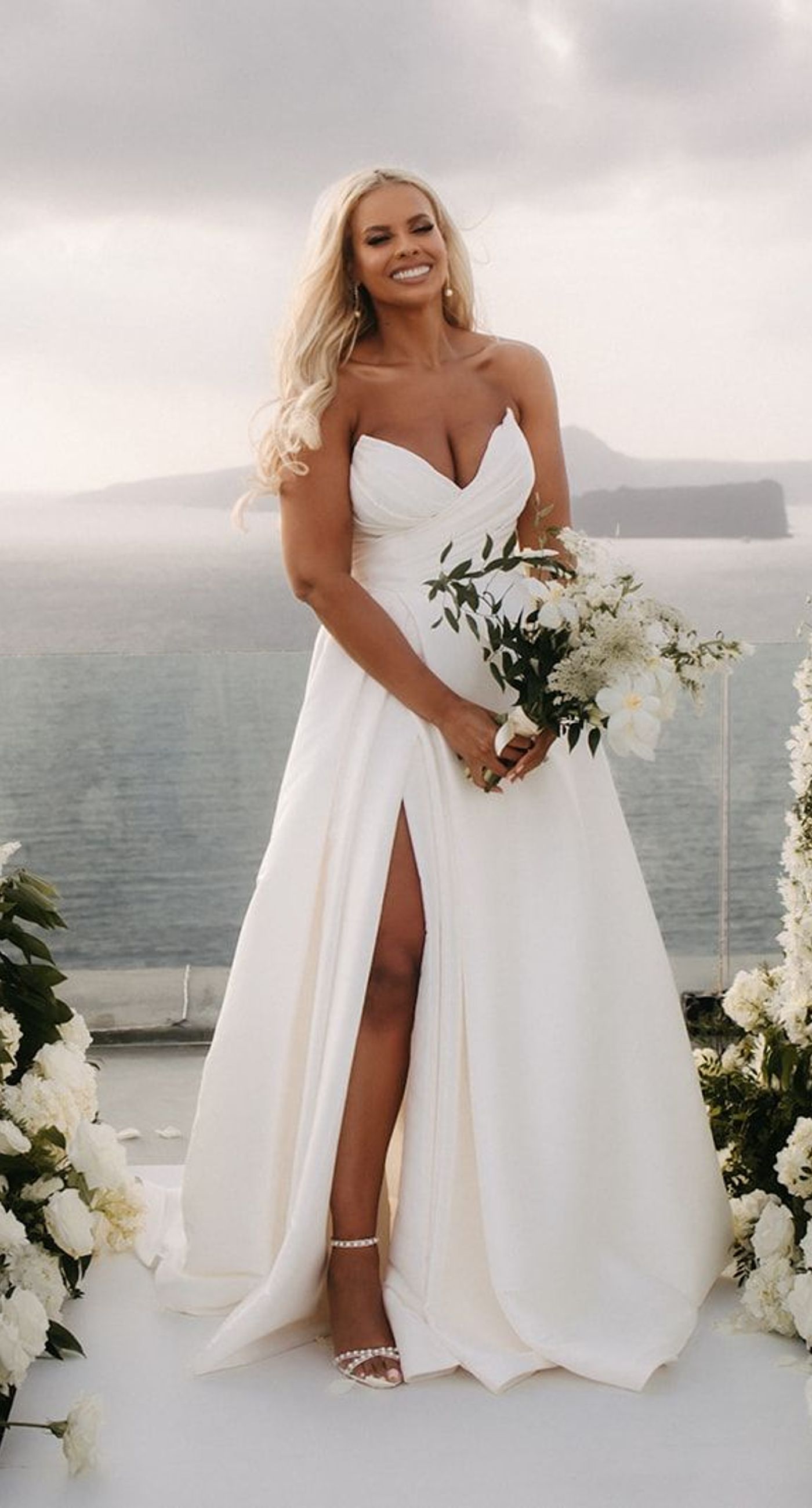 Lauren & William - Amidst a backdrop of oceanic vistas, the couple's radiant joy is accentuated by a floral pathway of elegant white blooms, creating an ethereal blend of romance and sophistication. The bride’s flowing gown mirrors the gentle waves beyond, while the groom’s crisp white suit with contrasting black accents adds a touch of modern elegance. The scene conveys a serene yet vibrant celebration, encapsulated by the natural light that washes over the space, infusing it with a warm, golden hue. This harmonious composition captures the essence of a perfect union, where nature and love intertwine seamlessly. - Photo 4