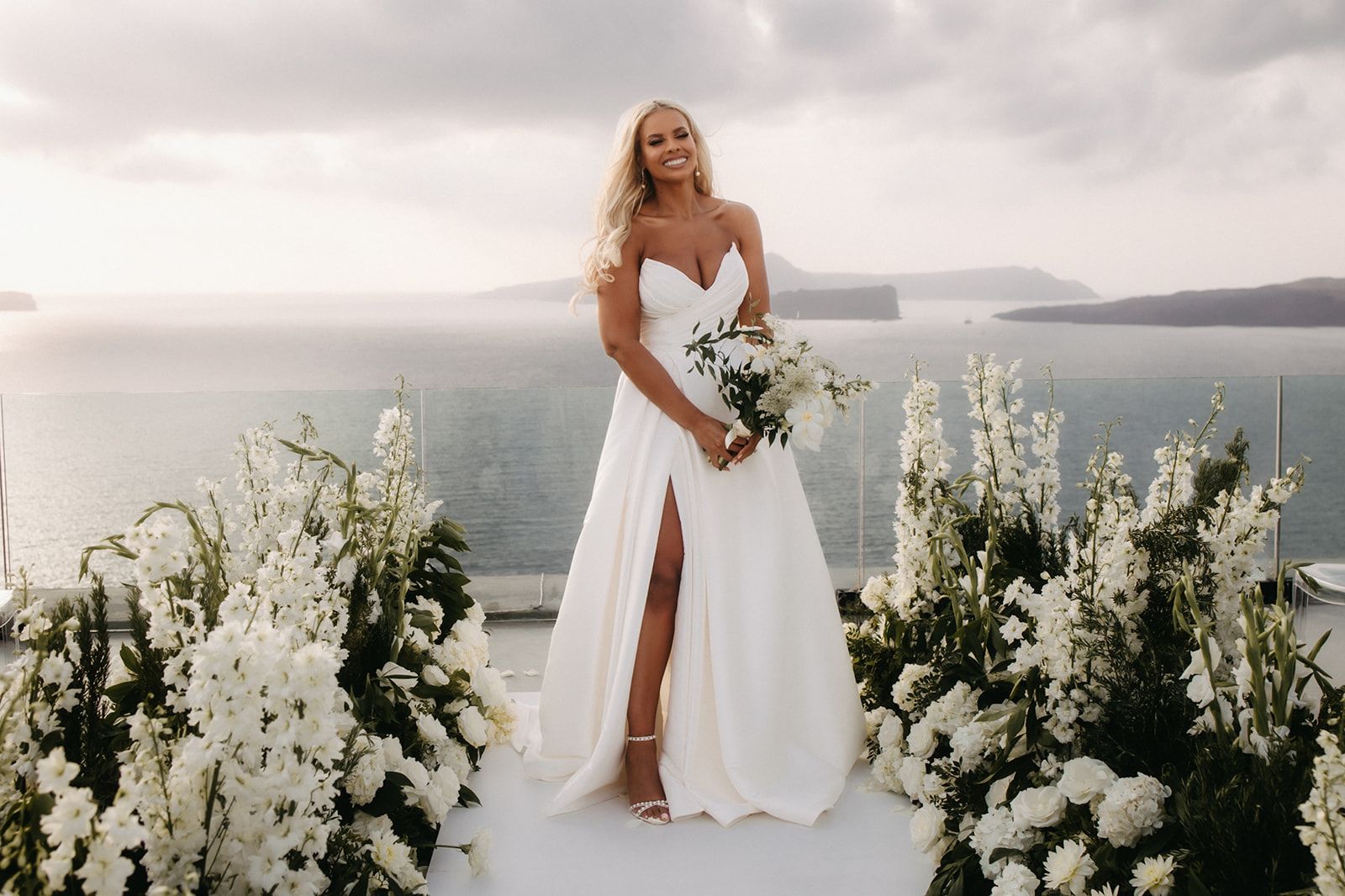 A bride in a strapless white satin gown with a deep sweetheart neckline and a high thigh slit stands smiling on a white aisle at Cavo Ventous in Greece. She holds a loose bouquet of white blooms and green foliage, and tall white delphinium and white roses line the aisle on both sides. Behind her, the sea and hazy grey islands stretch to the horizon under a soft, overcast sky, giving the scene a calm, airy feel.