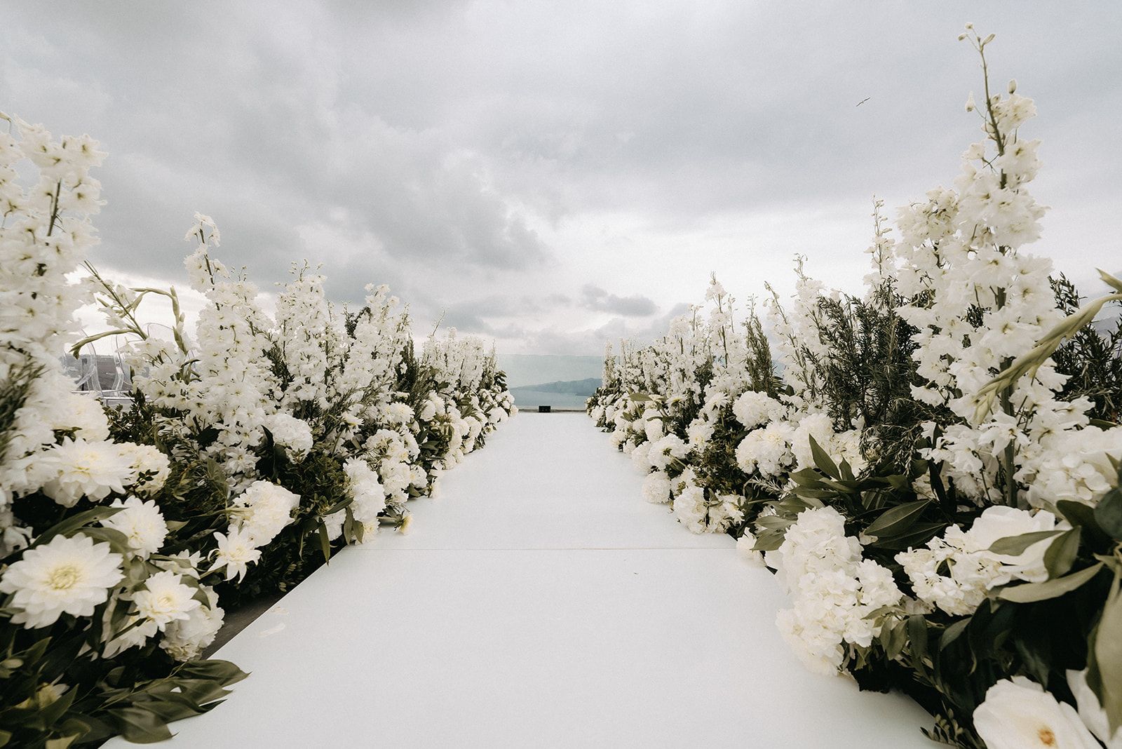 A white aisle runs straight toward the sea, lined on both sides with dense borders of white delphinium, white dahlias, hydrangeas and sprigs of green rosemary and olive foliage. These wedding flowers at Cavo Ventous in Greece are arranged in tall, lush rows beneath a soft, overcast grey sky. Distant hills fade into the water on the horizon, giving the scene a calm, monochrome and elegant feel.