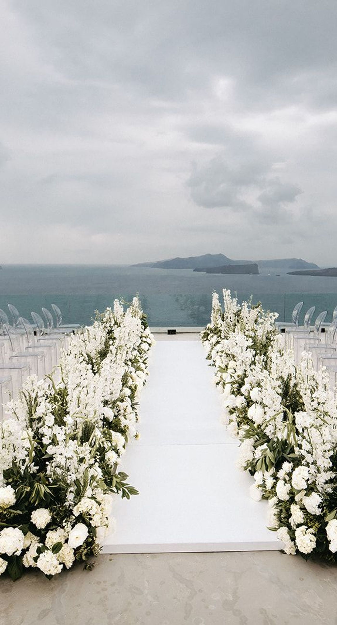 Lauren & William - Amidst a backdrop of oceanic vistas, the couple's radiant joy is accentuated by a floral pathway of elegant white blooms, creating an ethereal blend of romance and sophistication. The bride’s flowing gown mirrors the gentle waves beyond, while the groom’s crisp white suit with contrasting black accents adds a touch of modern elegance. The scene conveys a serene yet vibrant celebration, encapsulated by the natural light that washes over the space, infusing it with a warm, golden hue. This harmonious composition captures the essence of a perfect union, where nature and love intertwine seamlessly. - Photo 1