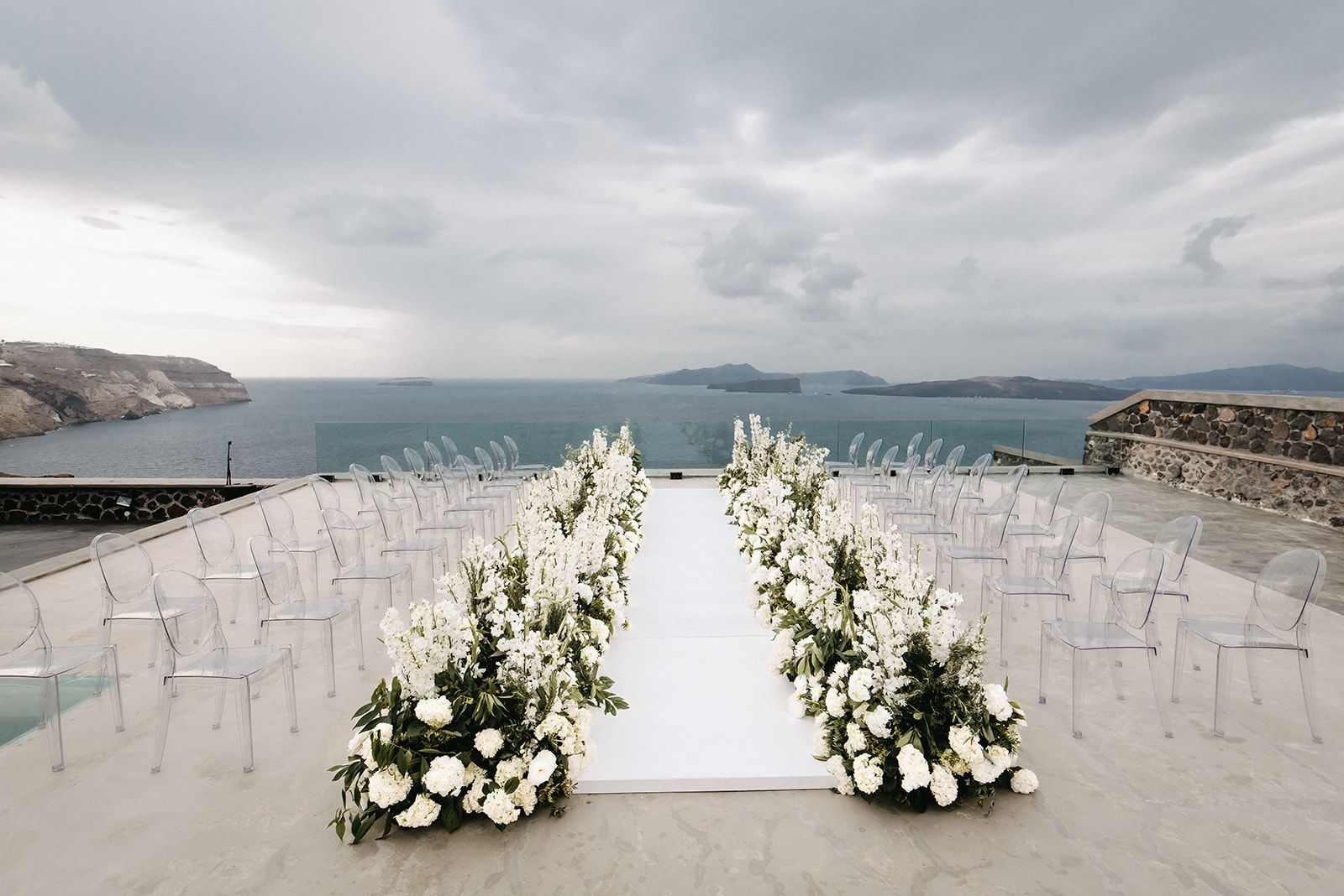 Two long borders of white flowers, mostly hydrangeas and white stock with sprigs of olive foliage, frame a white aisle runner leading down a stone terrace at Cavo Ventous, a wedding venue in Greece. Clear ghost chairs line both sides, looking out over a calm grey sea, with cliffs and small islands under a heavy, overcast sky. The muted palette of white, green and grey gives the wedding ceremony a serene, elegant mood.