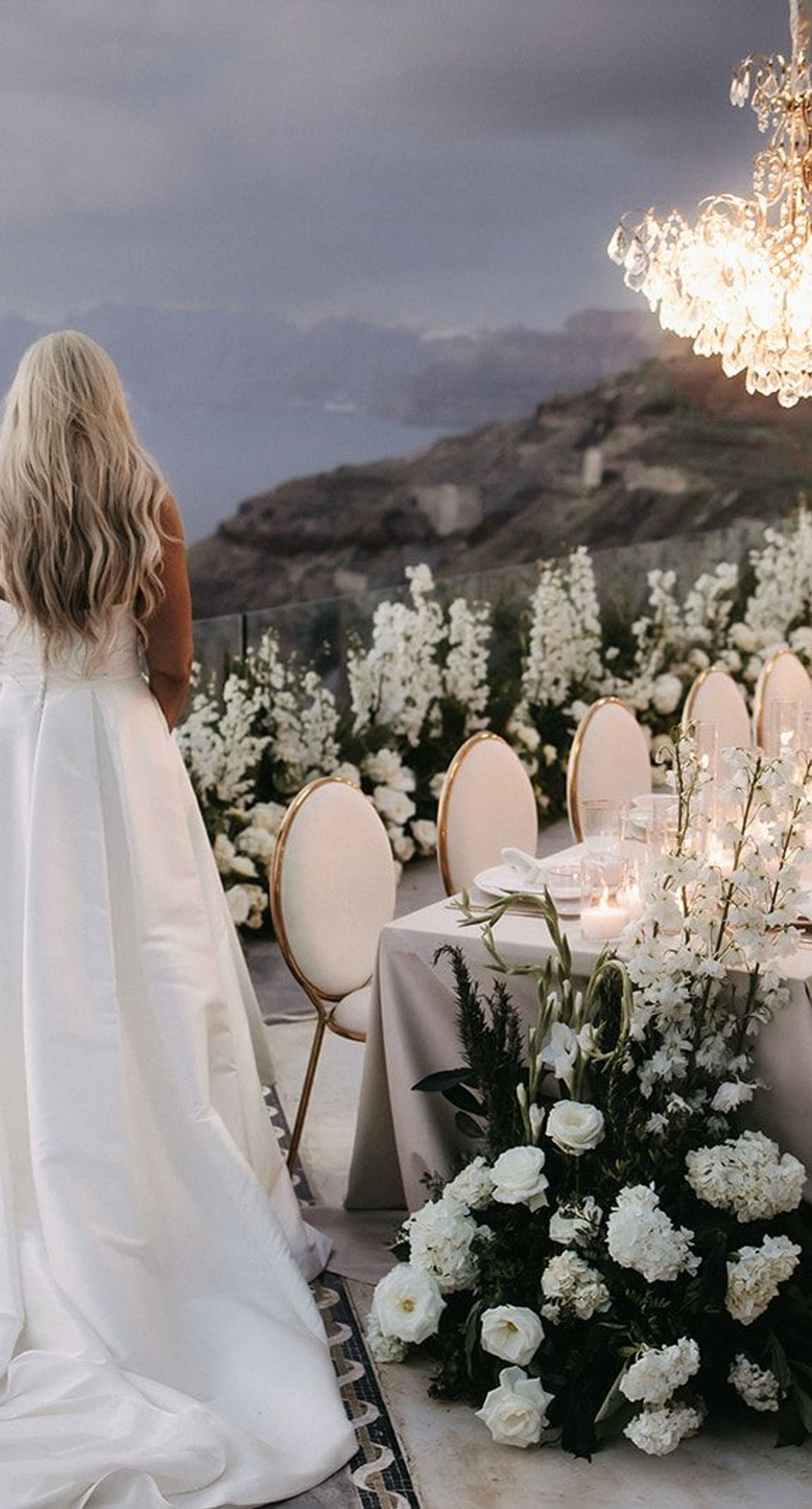 Lauren & William - Amidst a backdrop of oceanic vistas, the couple's radiant joy is accentuated by a floral pathway of elegant white blooms, creating an ethereal blend of romance and sophistication. The bride’s flowing gown mirrors the gentle waves beyond, while the groom’s crisp white suit with contrasting black accents adds a touch of modern elegance. The scene conveys a serene yet vibrant celebration, encapsulated by the natural light that washes over the space, infusing it with a warm, golden hue. This harmonious composition captures the essence of a perfect union, where nature and love intertwine seamlessly. - Photo 7