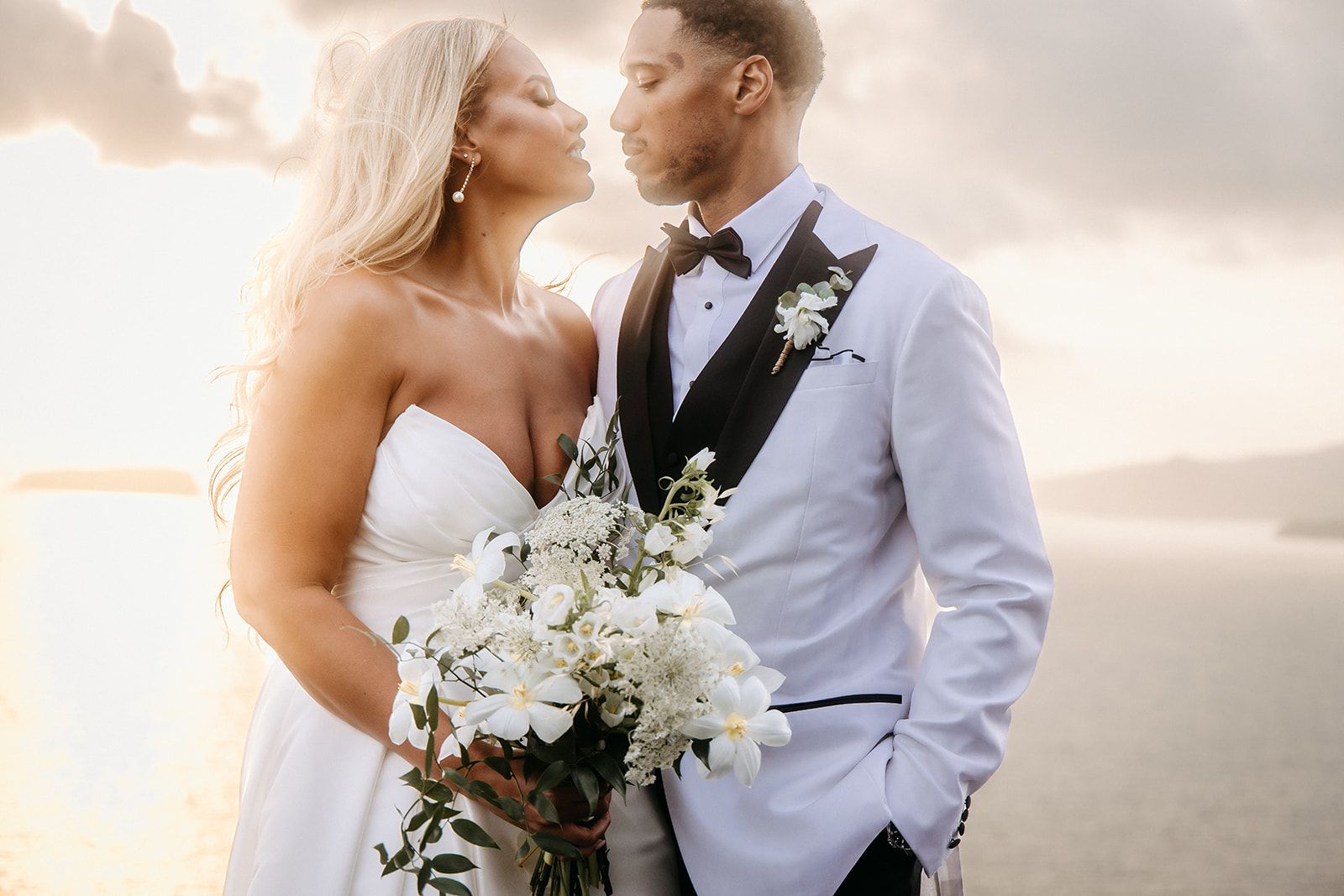 A bride in a white strapless gown and a groom in a white dinner jacket with a black satin lapel and bow tie lean in toward each other, eyes closed, as if about to kiss, against a soft, glowing sky at sunset. She holds a loose bouquet of white flowers, likely ranunculus and Queen Anne's lace, with trailing green foliage, while he wears a small white boutonniere on his lapel. This wedding flower arrangement was made for a wedding at Cavo Ventous in Greece.