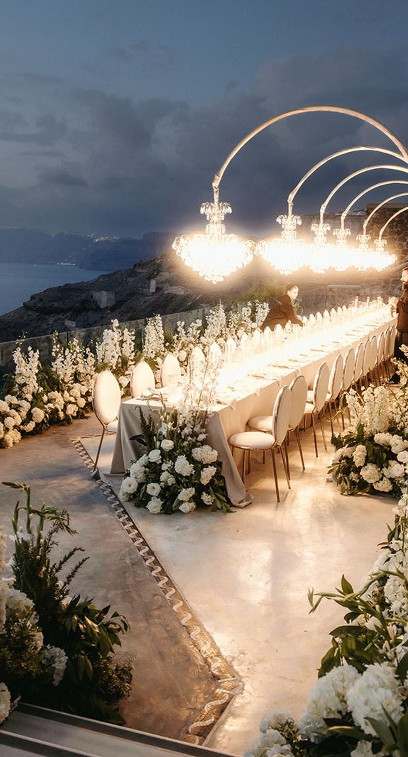 Lauren & William - Amidst a backdrop of oceanic vistas, the couple's radiant joy is accentuated by a floral pathway of elegant white blooms, creating an ethereal blend of romance and sophistication. The bride’s flowing gown mirrors the gentle waves beyond, while the groom’s crisp white suit with contrasting black accents adds a touch of modern elegance. The scene conveys a serene yet vibrant celebration, encapsulated by the natural light that washes over the space, infusing it with a warm, golden hue. This harmonious composition captures the essence of a perfect union, where nature and love intertwine seamlessly. - Photo 9