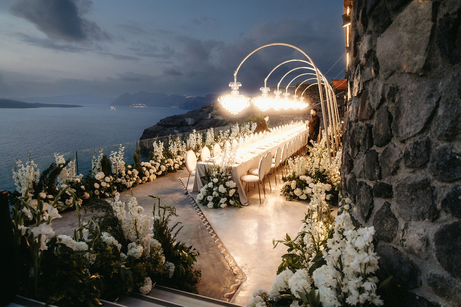 A long wedding dining table stretches along a stone terrace at dusk, set beneath a row of arched metal frames hung with glowing crystal chandeliers, with white delphinium, hydrangea and roses spilling along both edges. Cream upholstered chairs line the table, and the cliffside view over the calm blue sea at Cavo Ventous in Santorini, Greece, fades into the evening sky.