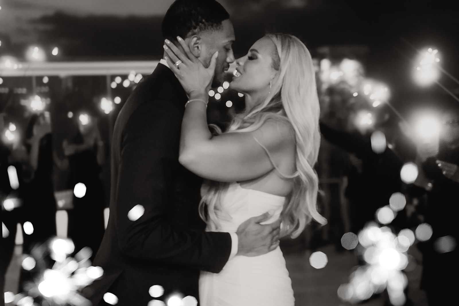 A black and white night-time photo of a bride in a white strapless gown and a groom in a dark suit sharing a tender kiss, her hands cradling his face and her long wavy hair falling down her back. Blurred sparkler lights glow around them, suggesting a wedding celebration at Cavo Ventous in Greece, where the couple's wedding flowers are arranged.