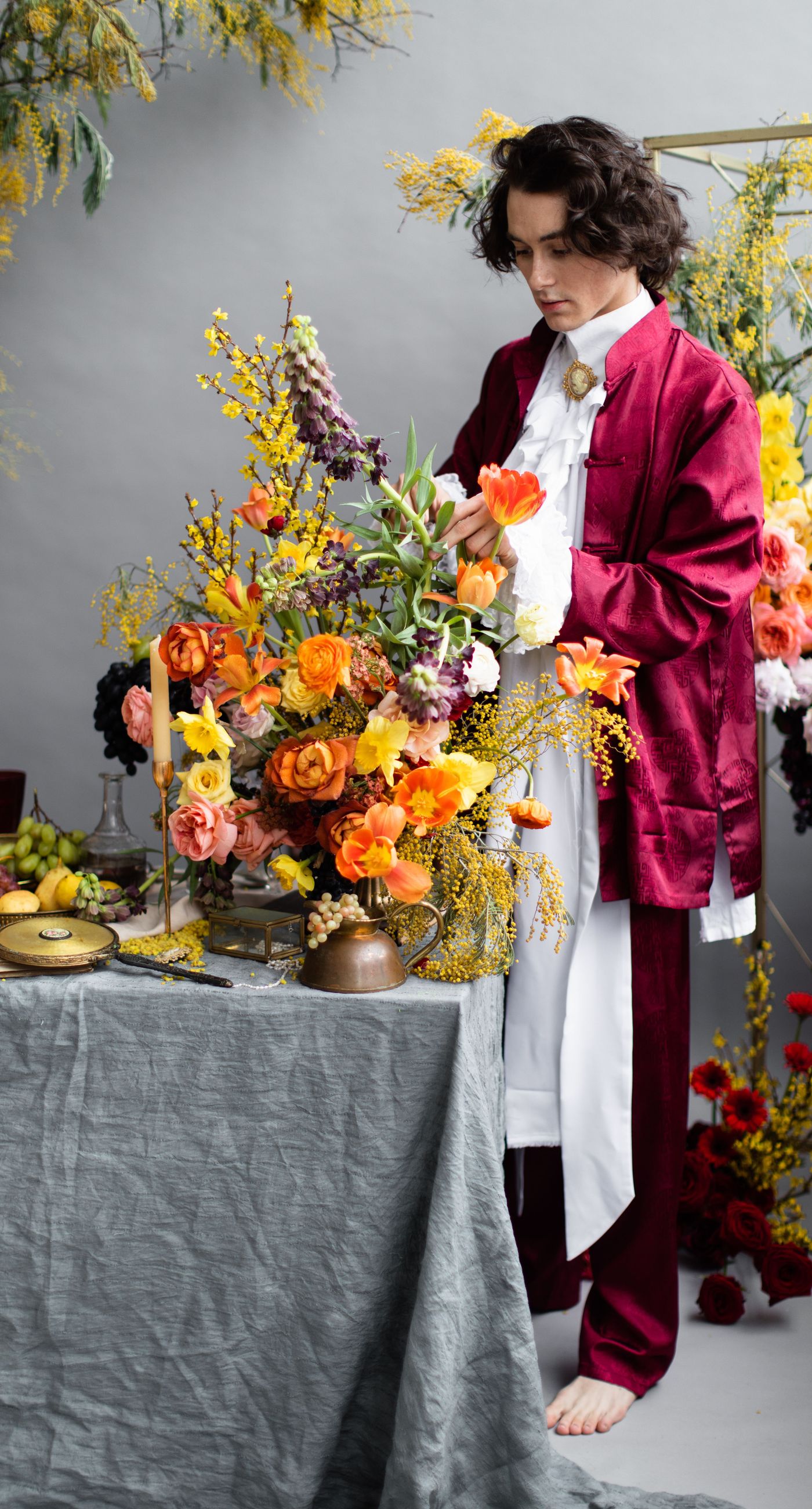 Medieval Dinner - In a symphony of colors, this floral arrangement weaves together vibrancy and elegance, creating an aesthetic that feels both timeless and contemporary. The vivid reds, yellows, and oranges of the blooms contrast strikingly against a soft, neutral backdrop, while a rich, wine-colored robe adds a touch of opulence. Surrounded by a lush abundance of fruits and elegant tableware, the setting evokes a sense of refined abundance, as if captured in a modern-day still life. The arrangement harmoniously balances the natural and the curated, transforming the space into a living work of art, and serving as a striking centerpiece for a florist's creative portfolio.
 - Photo 3