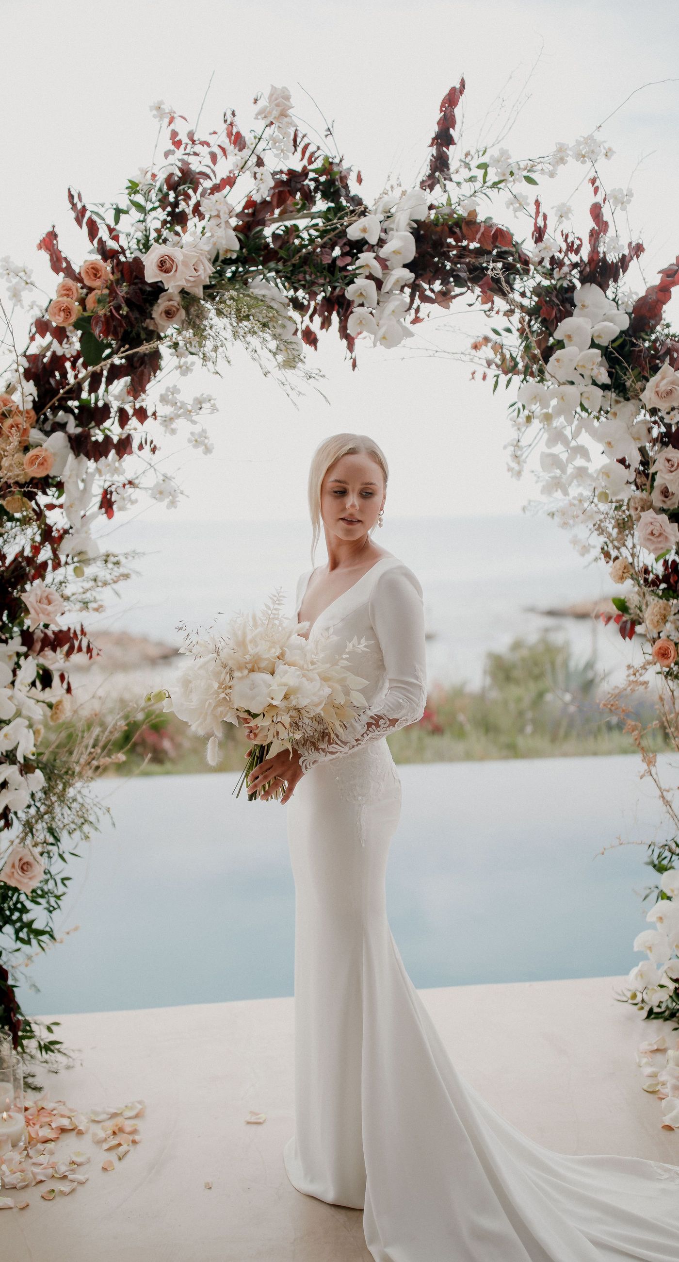 Monika & Ryan - Under a gentle sky that meets a serene coastline, a beautifully crafted floral arch frames an intimate moment of joy and elegance. Soft blush roses and pristine white orchids cascade amid deep burgundy leaves, creating a lush tapestry that echoes the surrounding natural beauty. The harmonious blend of textures and muted colors evokes a sense of timeless romance, making each element of the design feel both organic and artful. The scene captures the essence of a tranquil seaside celebration, where nature's breathtaking backdrop complements the exquisite floral artistry. - Photo 1