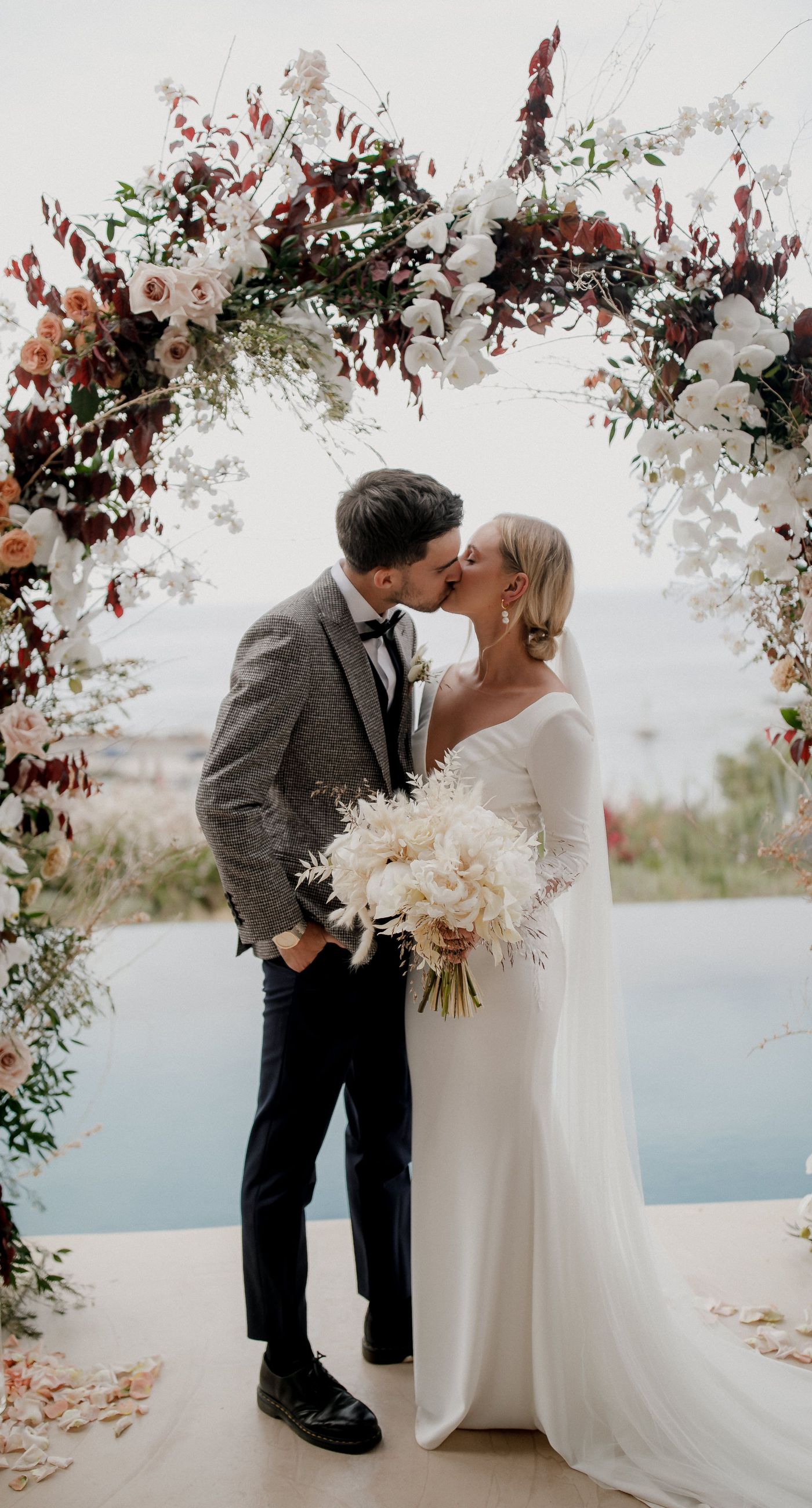 Monika & Ryan - Under a gentle sky that meets a serene coastline, a beautifully crafted floral arch frames an intimate moment of joy and elegance. Soft blush roses and pristine white orchids cascade amid deep burgundy leaves, creating a lush tapestry that echoes the surrounding natural beauty. The harmonious blend of textures and muted colors evokes a sense of timeless romance, making each element of the design feel both organic and artful. The scene captures the essence of a tranquil seaside celebration, where nature's breathtaking backdrop complements the exquisite floral artistry. - Photo 2