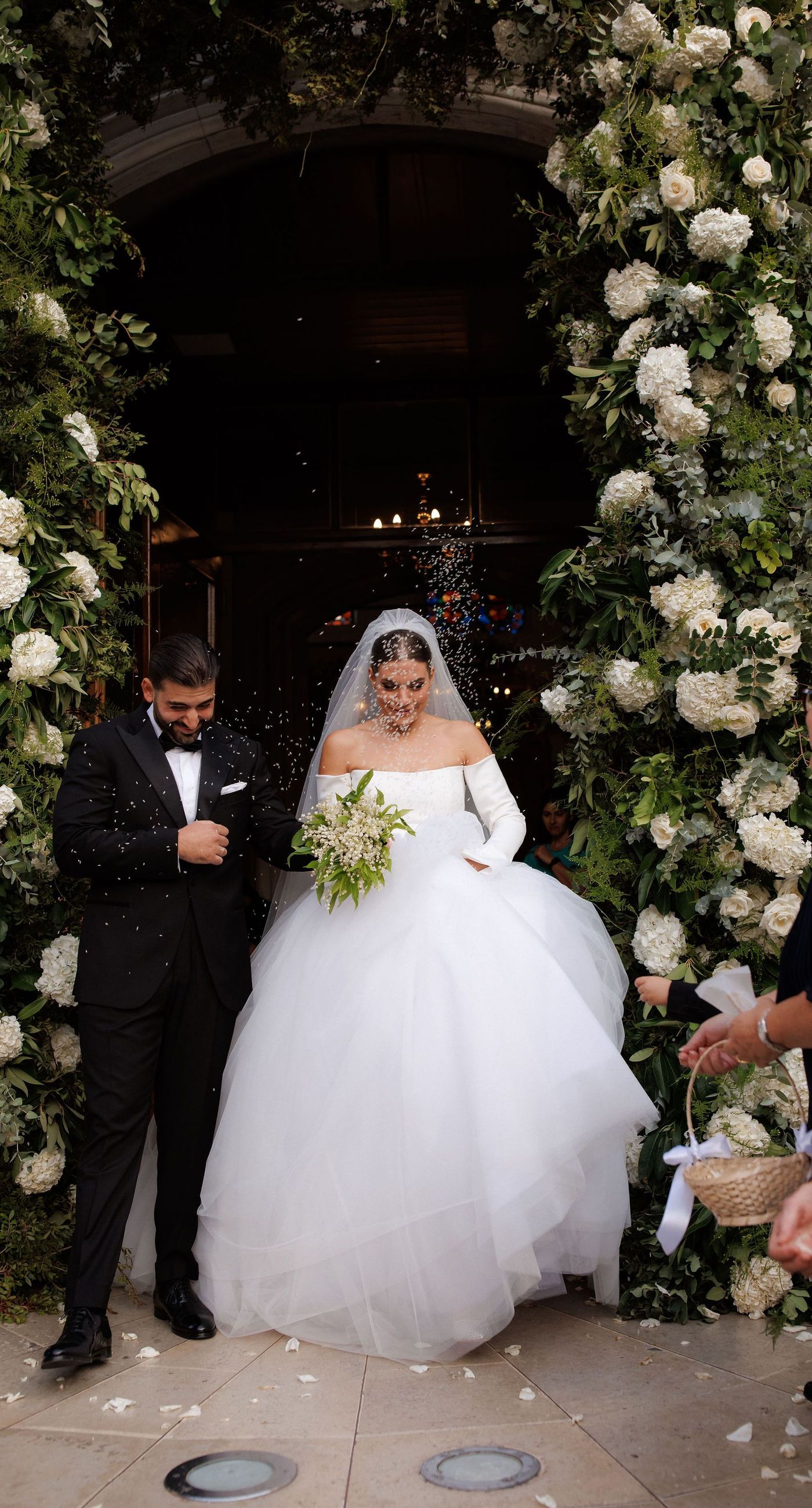Natalia & John  - Amidst a cascade of ivory roses and lush greenery, an elegantly composed scene unfolds, embodying a timeless romance. The bride, draped in a voluminous white gown, holds a delicate bouquet of blooms that mirrors the floral splendor surrounding her. The groom, in classic black tie, complements the serene palette with a touch of timeless elegance. This artistic composition brings together the soft whispers of petals and the gentle embrace of forgotten days, creating a vibrant harmony between nature's charm and heartfelt human moments. The florals, artfully arranged, create an enchanting backdrop that speaks of new beginnings and enduring beauty. - Photo 2