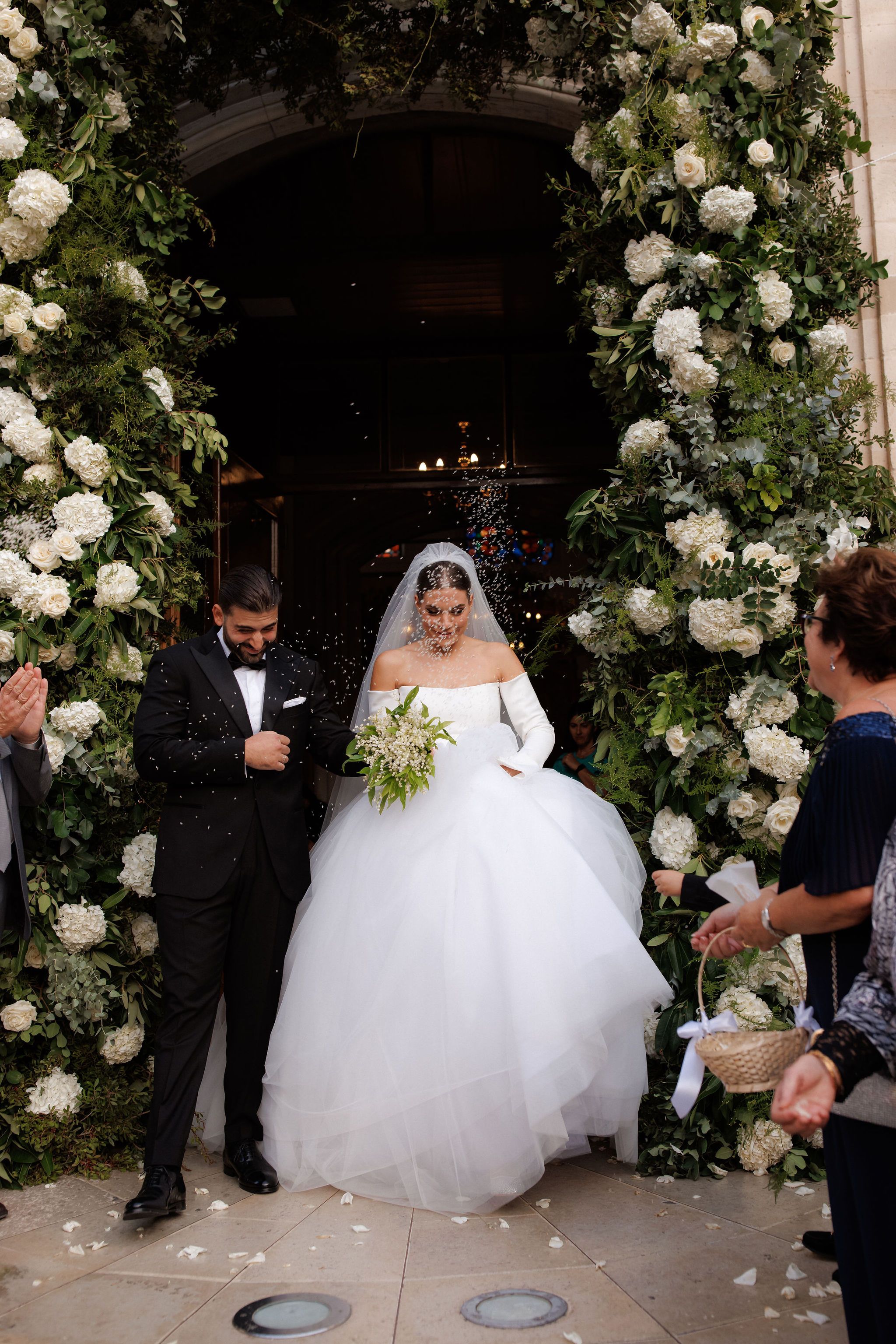 A bride and groom step out through a church doorway framed by tall arches of white hydrangeas, white roses and silver-green eucalyptus, all part of the wedding flowers for this Greece celebration. Confetti drifts through the air around them as they walk forward, the bride in a voluminous white ballgown with long gloves and a veil, holding a small bouquet of white blooms and greenery, while the groom in a black tuxedo smiles and looks down. Guests on the right watch and applaud, and the dim interior behind them glows with candlelight.