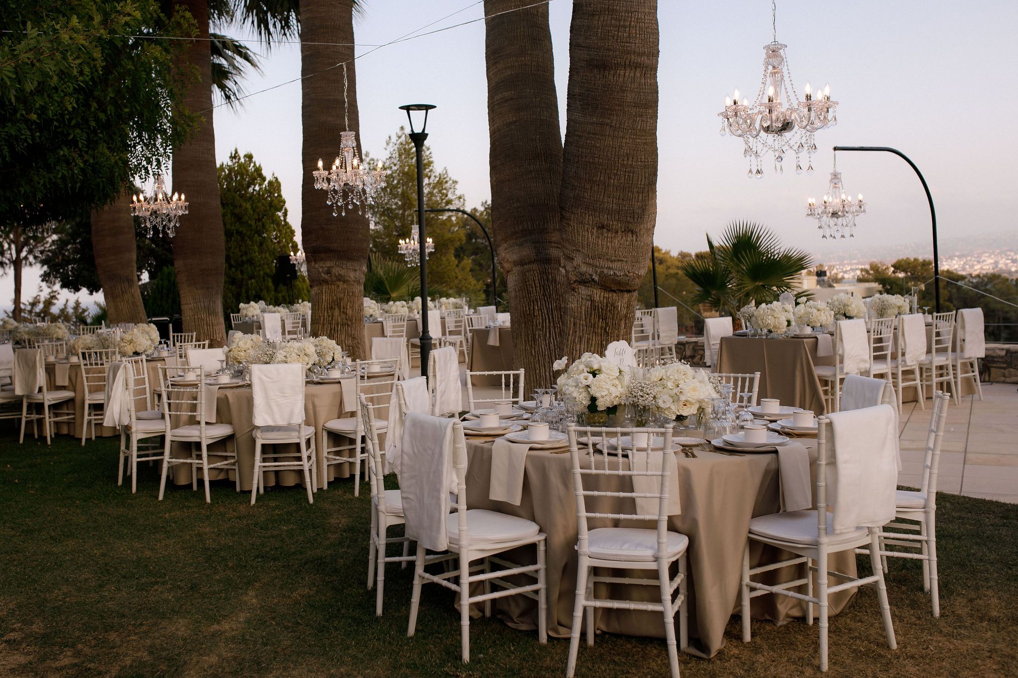 Round tables dressed in taupe linen sit on a lawn beneath tall palm trunks, with white Chiavari chairs draped in ivory covers. Centrepieces of white hydrangeas, white roses and baby's breath rise above the place settings, and crystal chandeliers hang among the palms at dusk. This is an outdoor wedding reception in Greece, styled in soft neutrals with a calm, elegant mood.