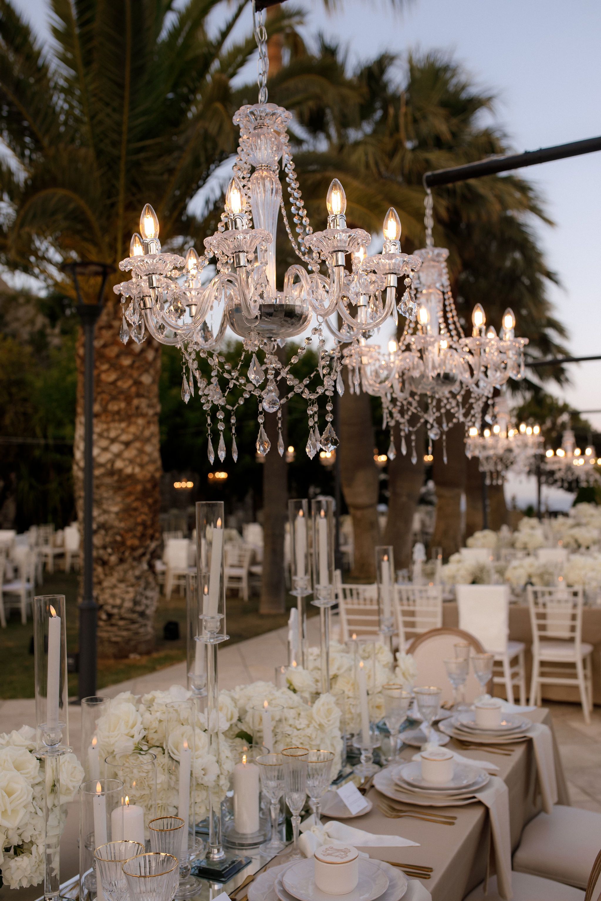 Crystal chandeliers with dangling beaded drops hang at different heights above an outdoor wedding reception in Greece, their candle-style bulbs glowing warmly against the dusk sky. Tall palm trunks rise behind them, and below, clear glass hurricane vases hold white pillar candles, with white flowers and white chairs visible among the tables in the soft blur beyond.