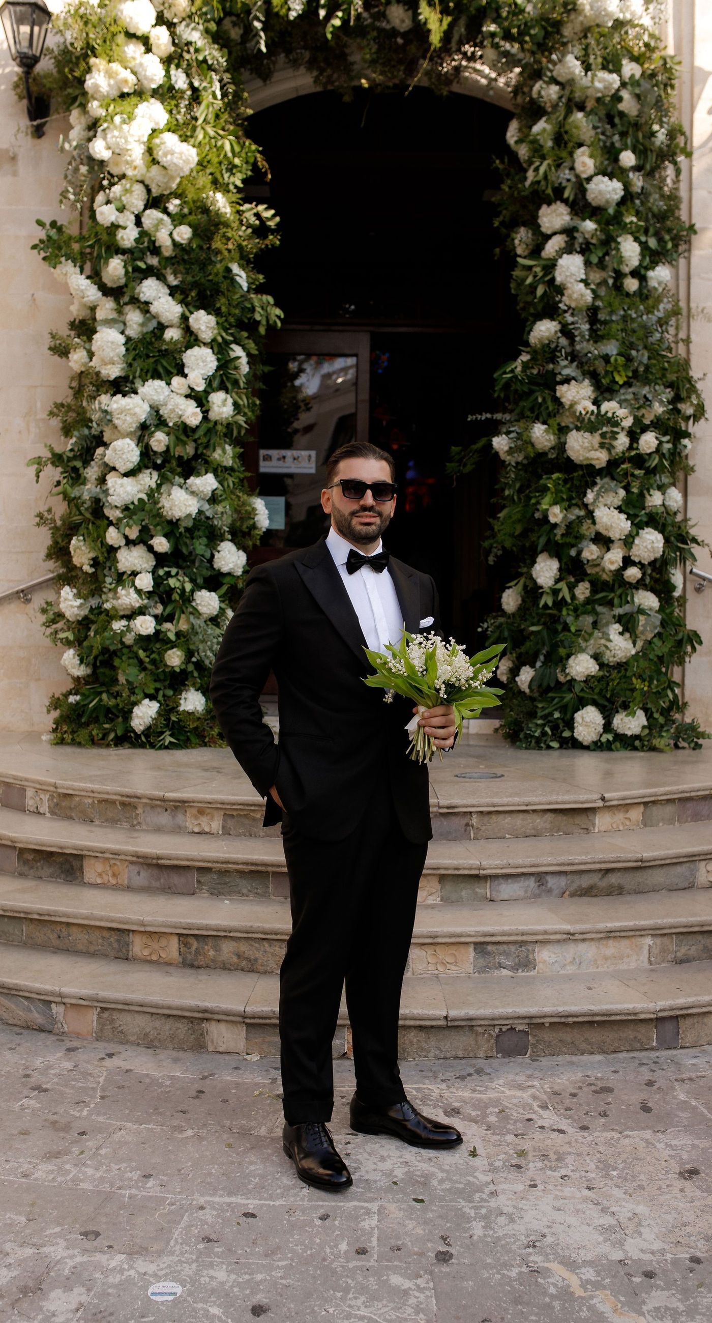 Natalia & John  - Amidst a cascade of ivory roses and lush greenery, an elegantly composed scene unfolds, embodying a timeless romance. The bride, draped in a voluminous white gown, holds a delicate bouquet of blooms that mirrors the floral splendor surrounding her. The groom, in classic black tie, complements the serene palette with a touch of timeless elegance. This artistic composition brings together the soft whispers of petals and the gentle embrace of forgotten days, creating a vibrant harmony between nature's charm and heartfelt human moments. The florals, artfully arranged, create an enchanting backdrop that speaks of new beginnings and enduring beauty. - Photo 1