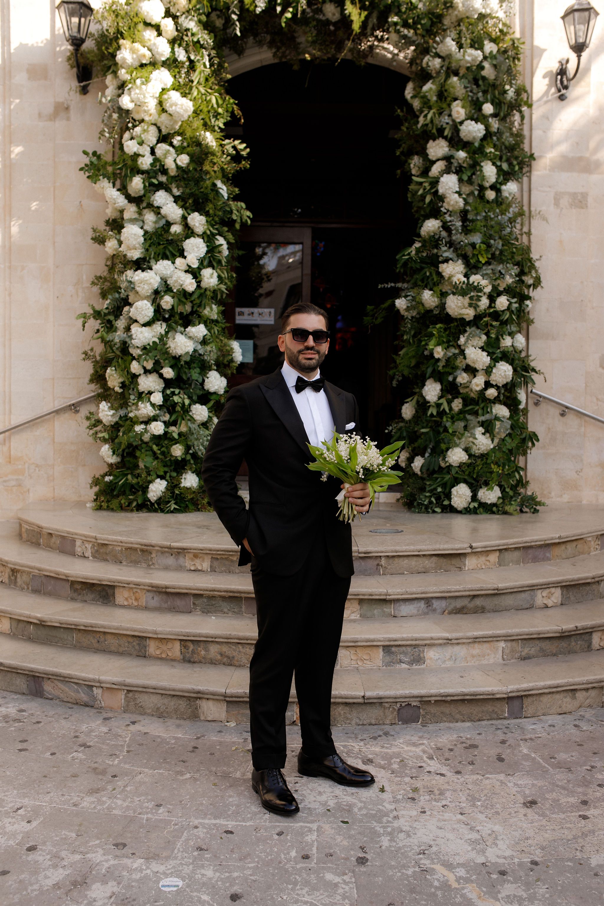 A groom in a black tuxedo, bow tie and dark sunglasses stands on pale stone steps, one hand in his pocket and a small bouquet of lily of the valley in the other. Behind him, two tall columns of white hydrangeas, white garden roses and green eucalyptus wrap up either side of a church doorway, forming a lush wedding arch. This is a wedding flower installation in Greece, with a crisp, elegant all-white and green palette.